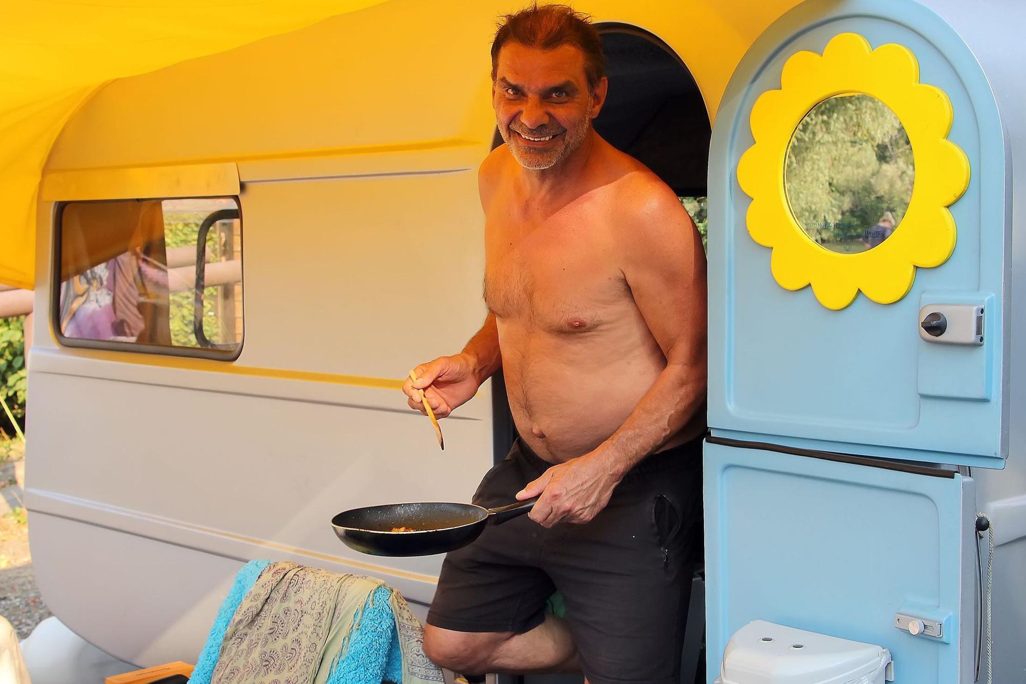 Das Bild zeigt einen Mann mit Pfanne in der Hand an seinem Campingwagen.
