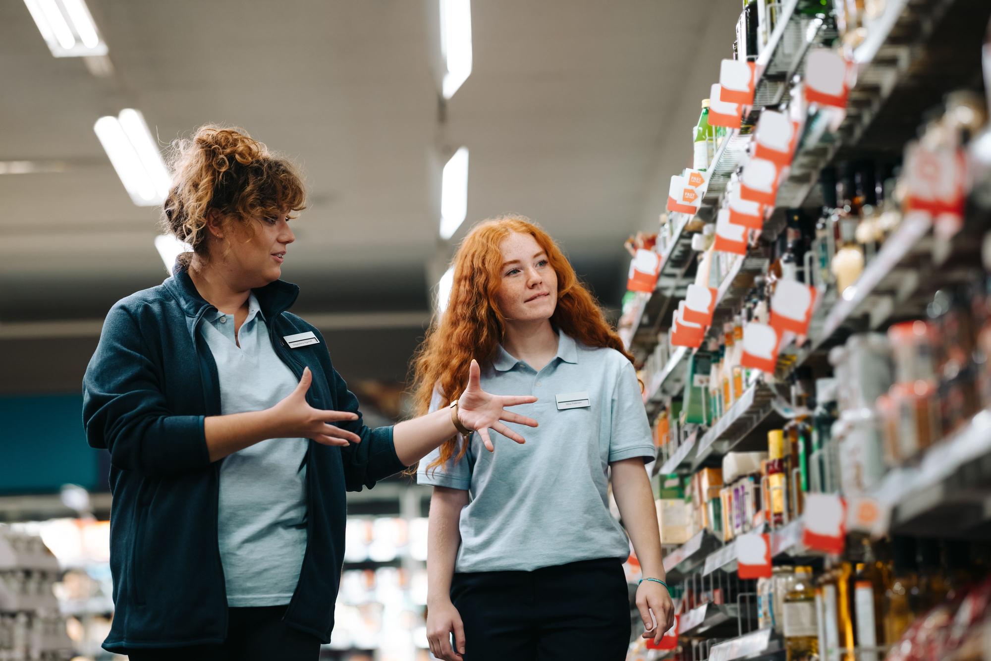 Zwei Frauen in Arbeitskleidung stehen in einem Supermarkt zwischen Regalen. Es wirkt wie eine Einarbeitung oder Schulung im Einzelhandel.
