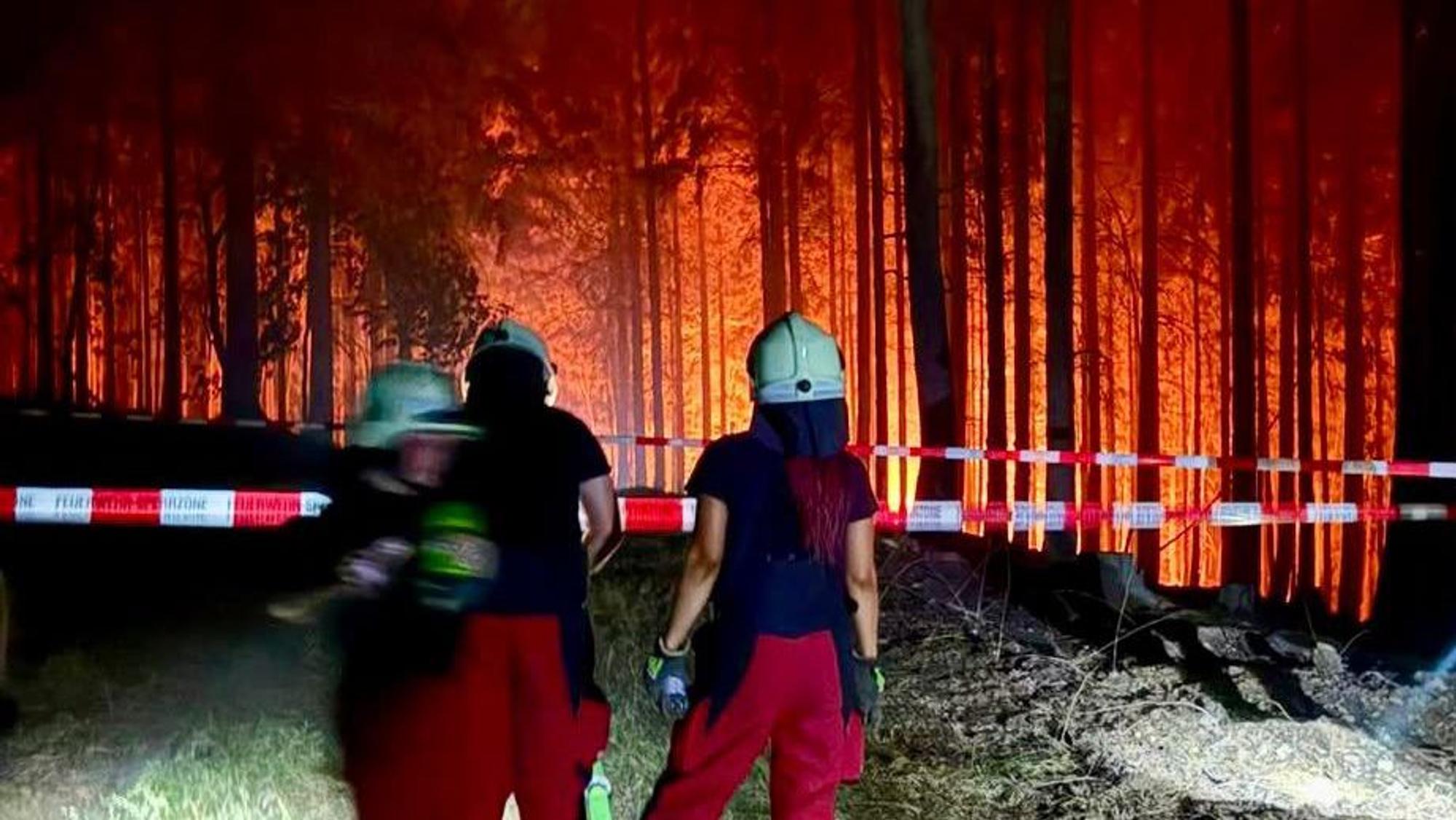 Feuerwehrleute aus Overath stehen an einer Absperrung vor einem brennenden Wald bei Düren.