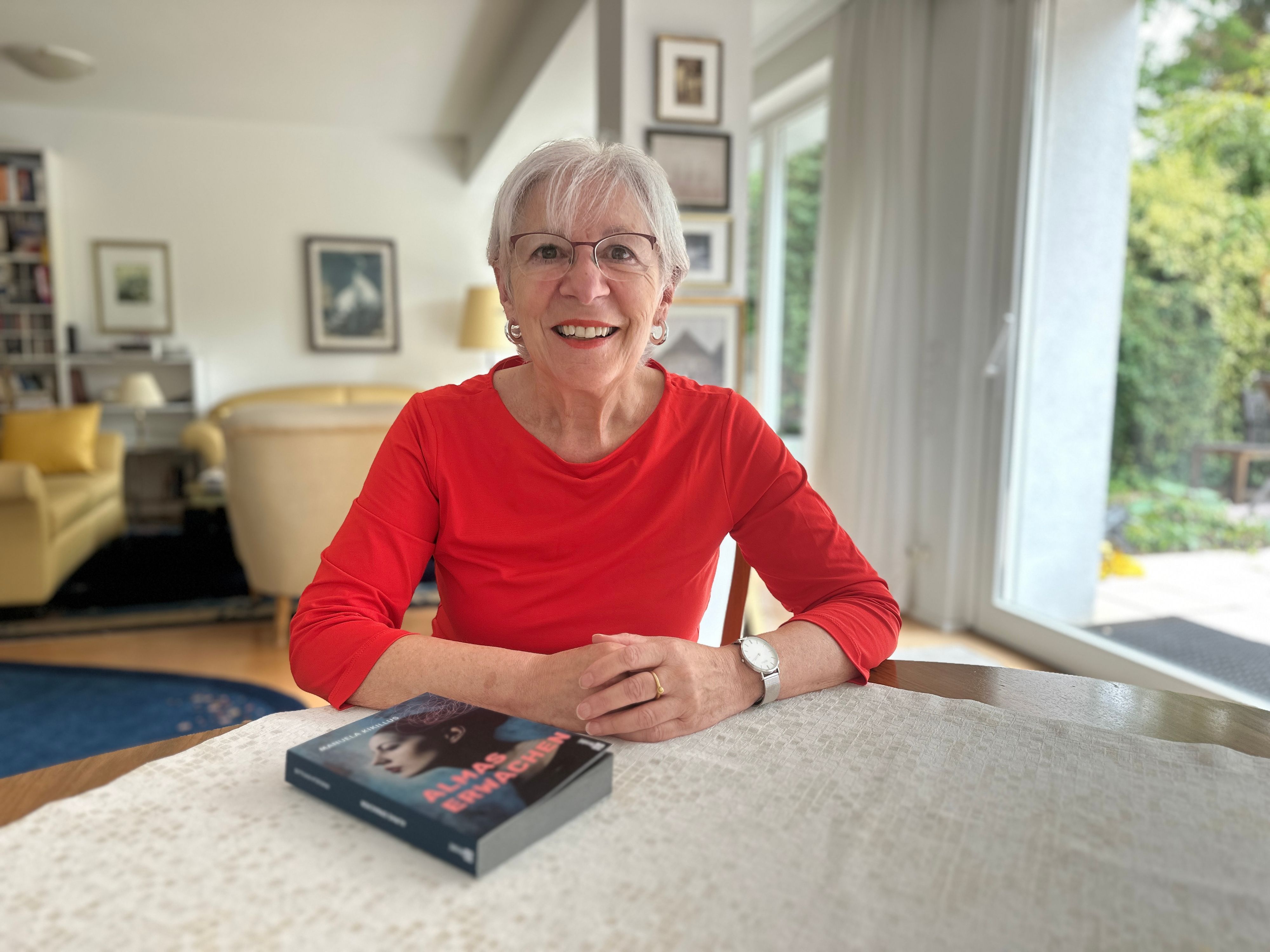 Eine Frau mit weißen, kurzen Haaren und rotem Oberteil, sitzt an einem Tisch. Vor ihr liegt ein Buch mit dem Titel Almas Erwachen.
