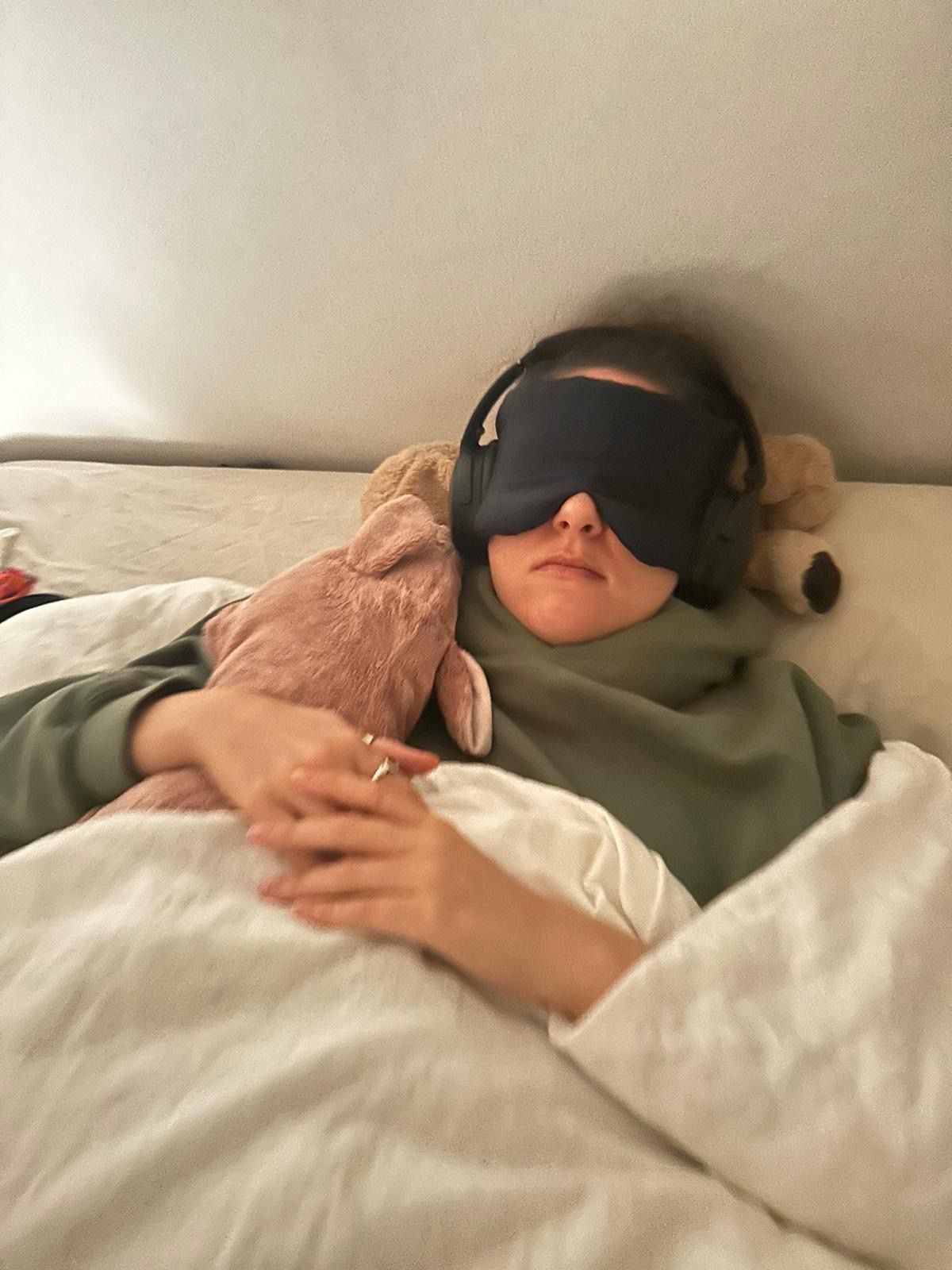 Eine Frau liegt mit Schlafmasken und Kopfhörern im Bett.