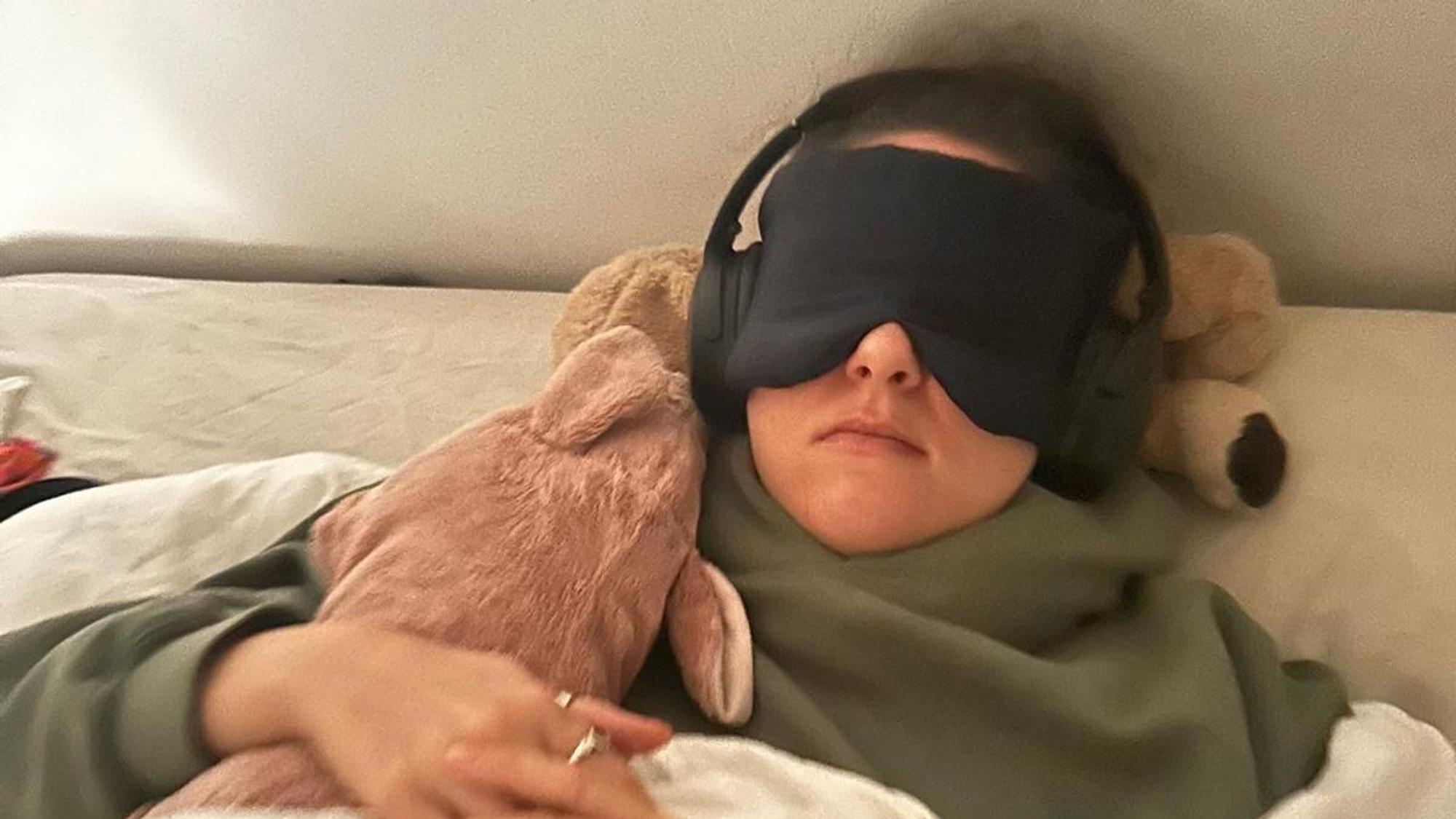 Eine Frau liegt mit Schlafmasken und Kopfhörern im Bett.