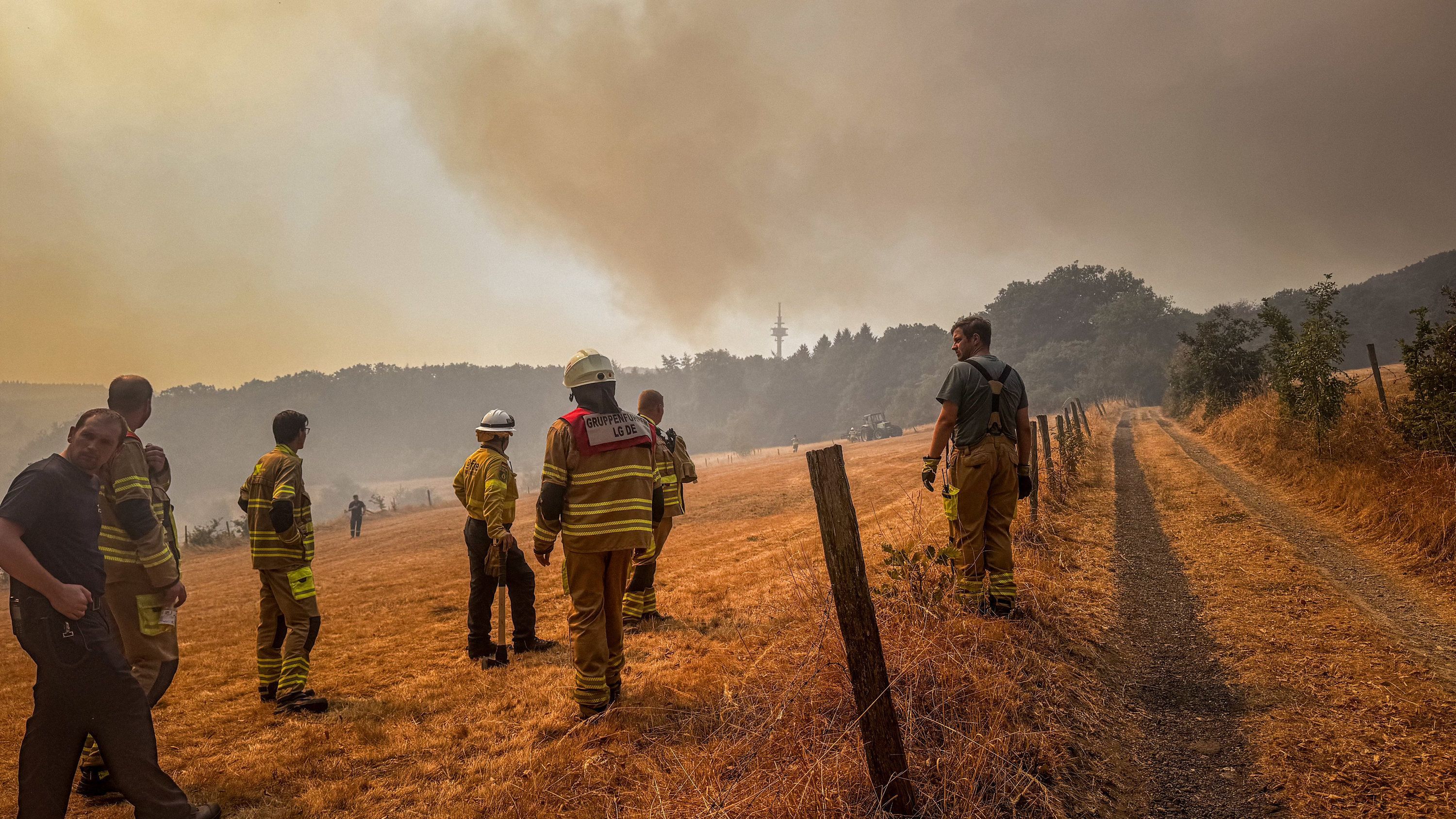 Verschiedene Einsatzkräfte stehen am 14. August 2026 auf einem Feld Nahe Hürtgenwald und blicken in Richtung eines Waldbrands, der Himmel ist rot gefärbt und durch Rauch verdunkelt. 

Zum KNA-Interview mit Bischof Helmut Dieser „Im Hürtgenwald war das ganze Land für die Menschen da“