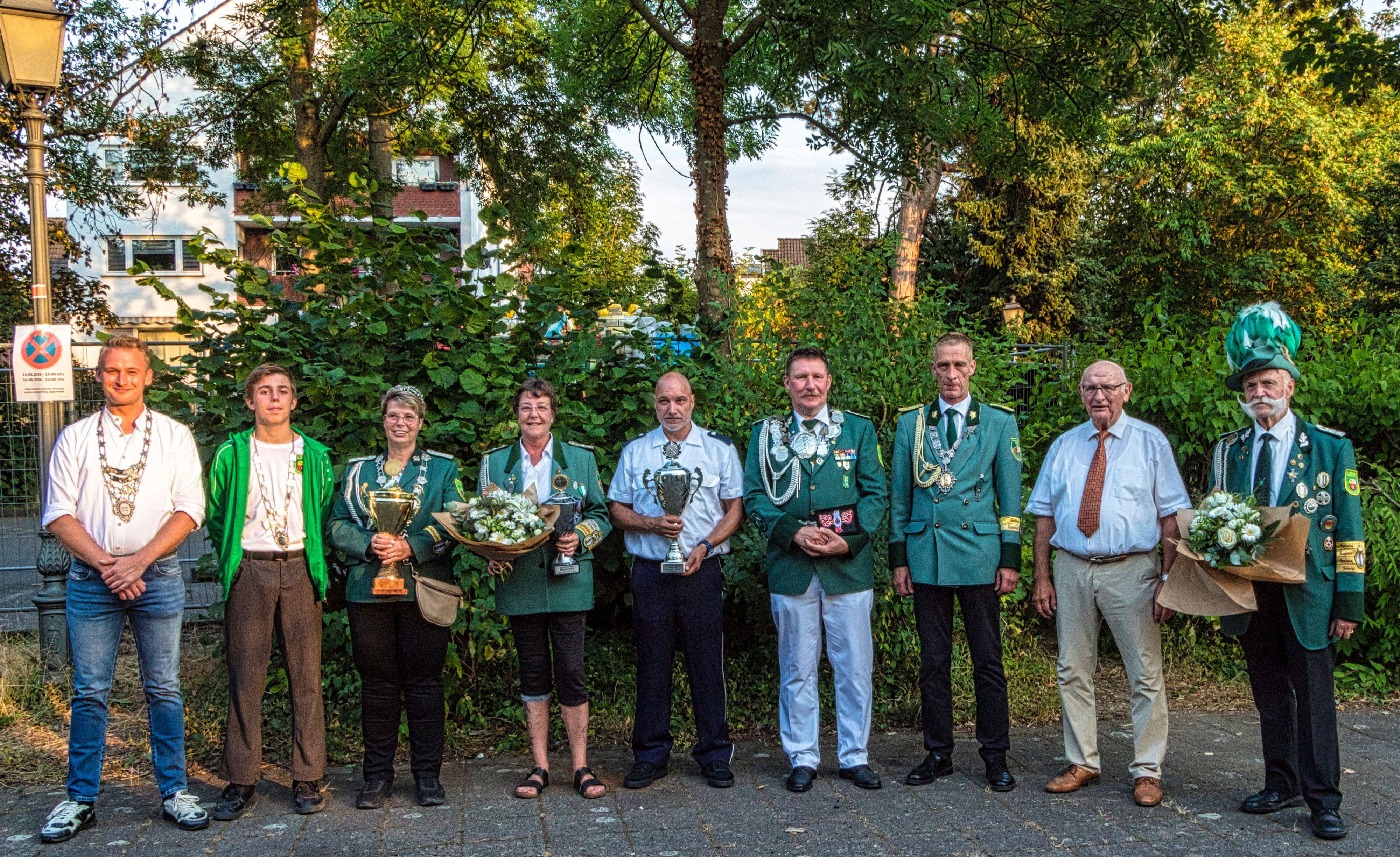 Neun Männer und Frauen, zum Teil in Uniform, stehen nebeneinander. Einige halten Pokale oder Blumensträuße in die Kamera.
