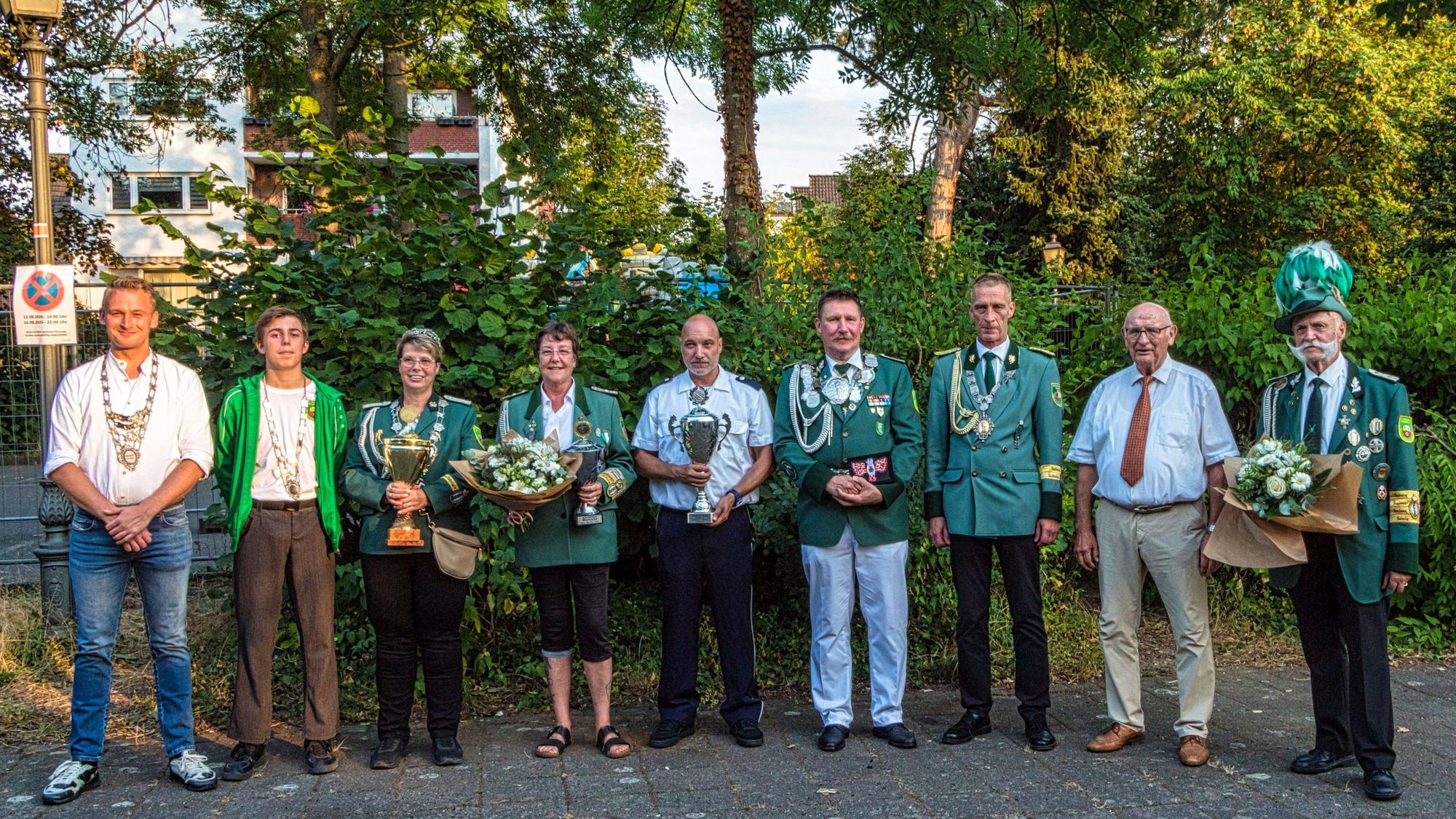Neun Männer und Frauen, zum Teil in Uniform, stehen nebeneinander. Einige halten Pokale oder Blumensträuße in die Kamera.