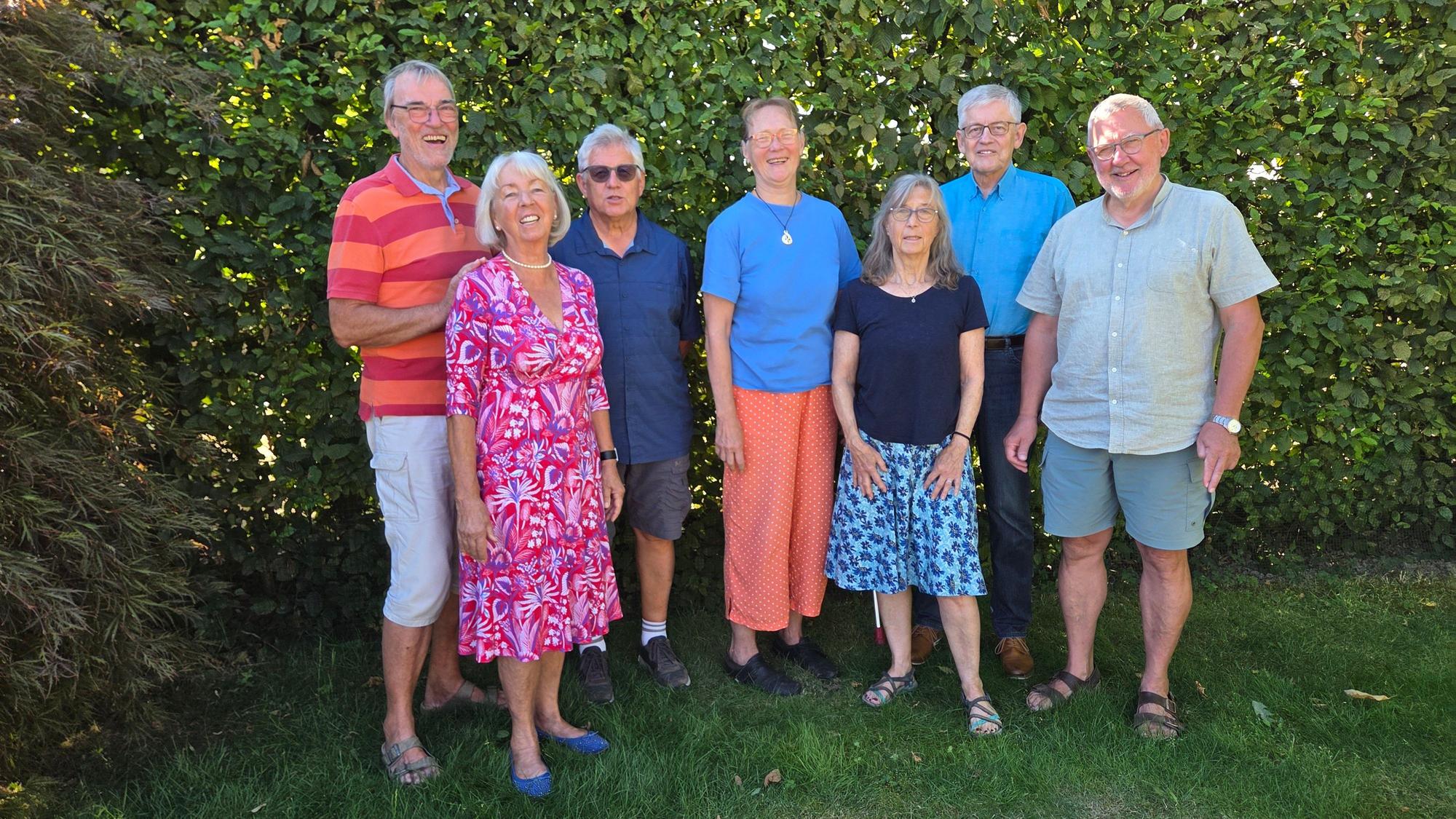 Eine Gruppe gut gelaunter Menschen steht vor einer Hecke auf einem Rasen und lächelt.