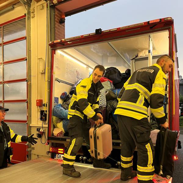 Zwei Feuerwehrmänner laden Reisegepäck aus einem Feuerwehrfahrzeug aus.