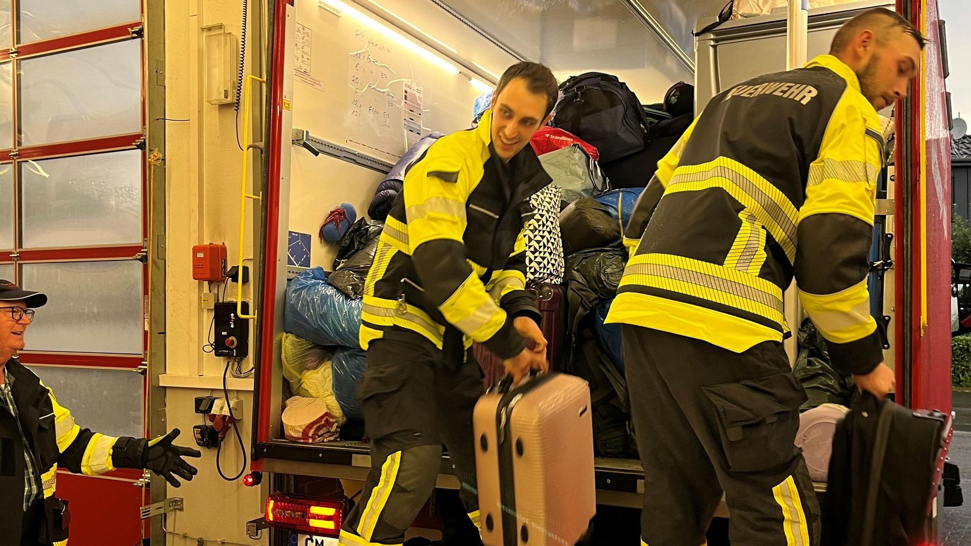 Zwei Feuerwehrmänner laden Reisegepäck aus einem Feuerwehrfahrzeug aus.