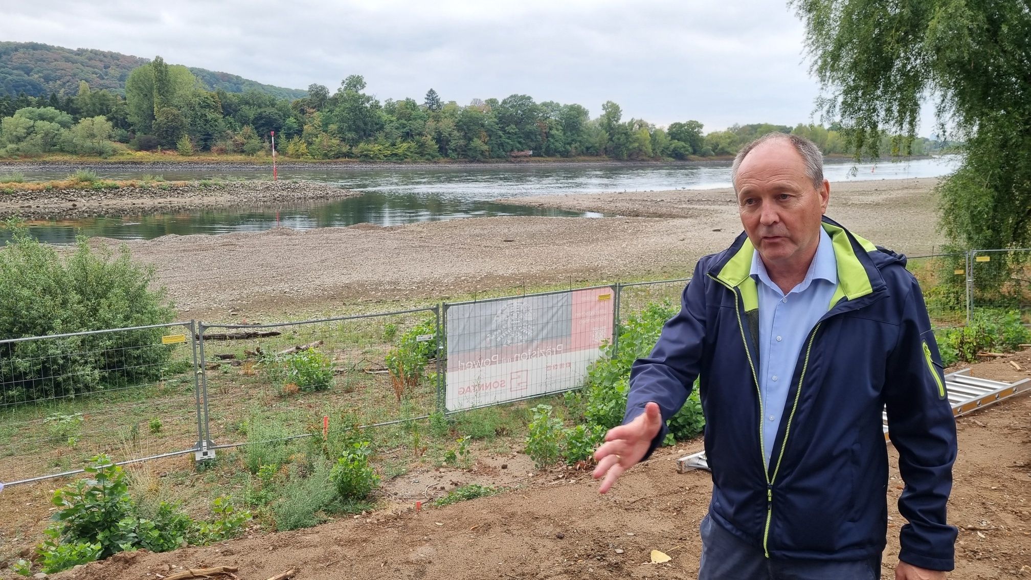 Ein Mann mit blauer Jacke steht neben einem Bauzaun am Ufer des Rheins.