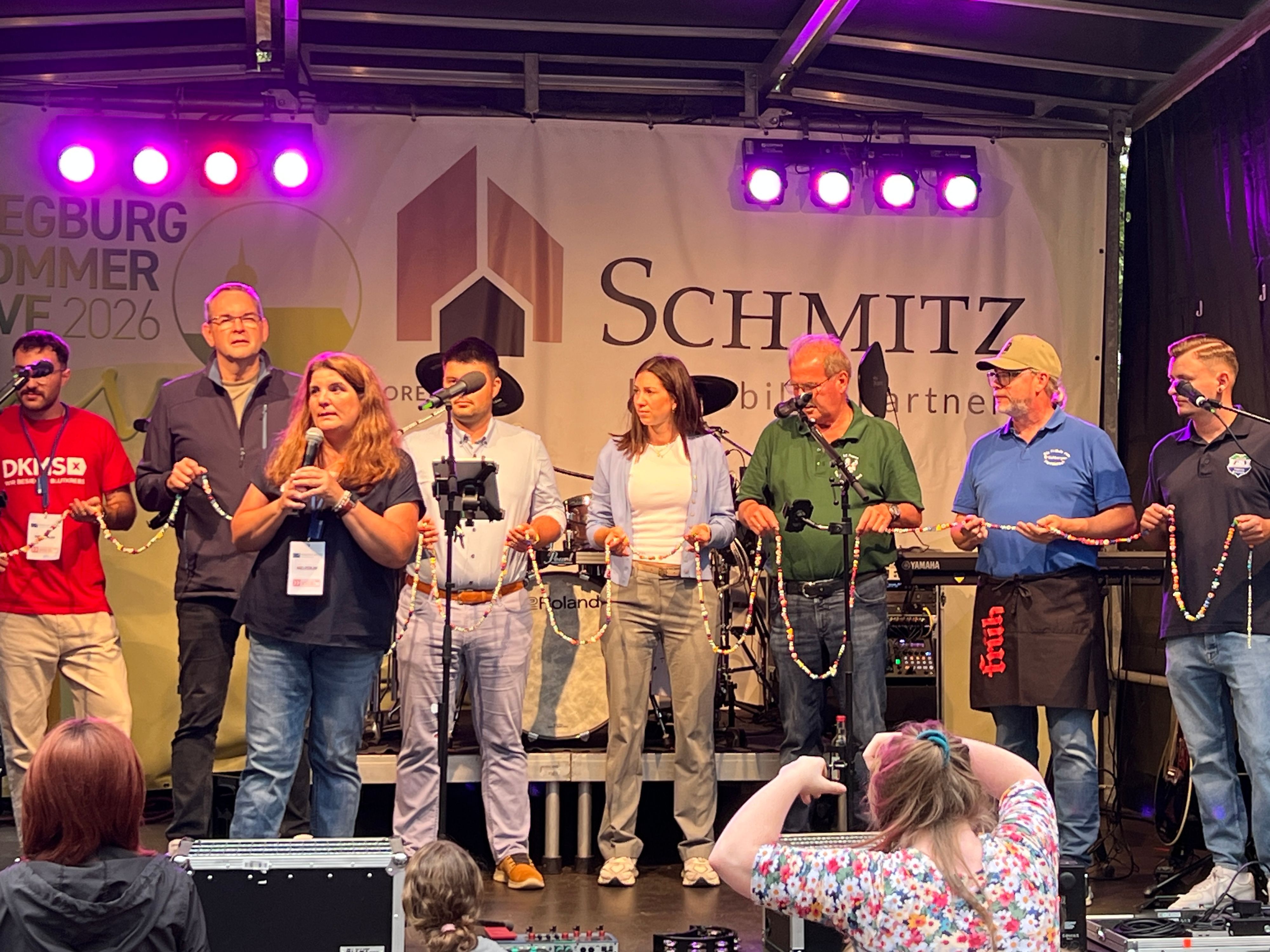 Auf dem Bild halten Helfende, darunter BG-Vorsitzende Sandra Weinert (mit Mikro) und Bürgermeister Stefan Rosemann (links dahinter), die „Mutmach-Kette“ von Max in den Händen.
