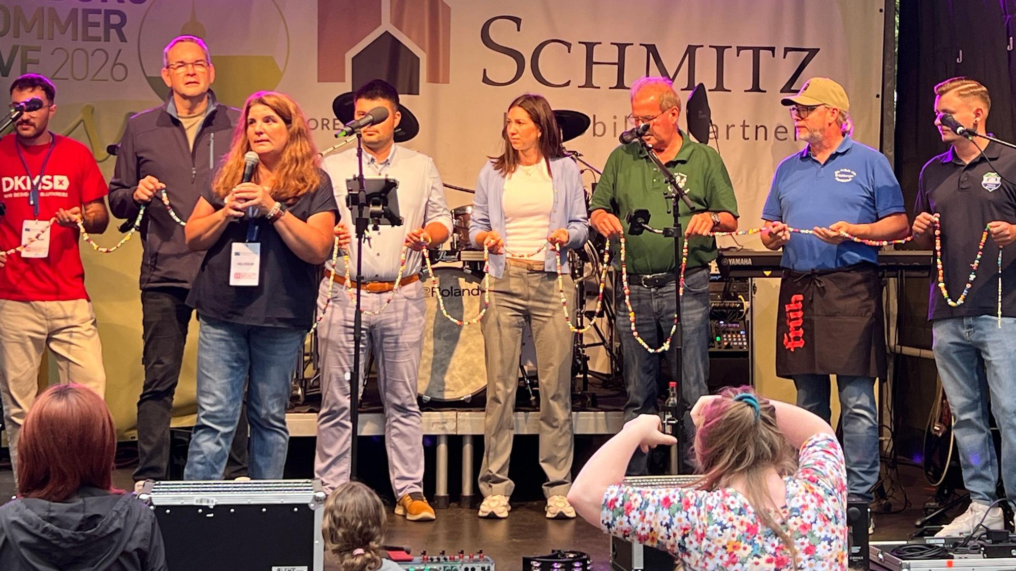Auf dem Bild halten Helfende, darunter BG-Vorsitzende Sandra Weinert (mit Mikro) und Bürgermeister Stefan Rosemann (links dahinter), die „Mutmach-Kette“ von Max in den Händen.