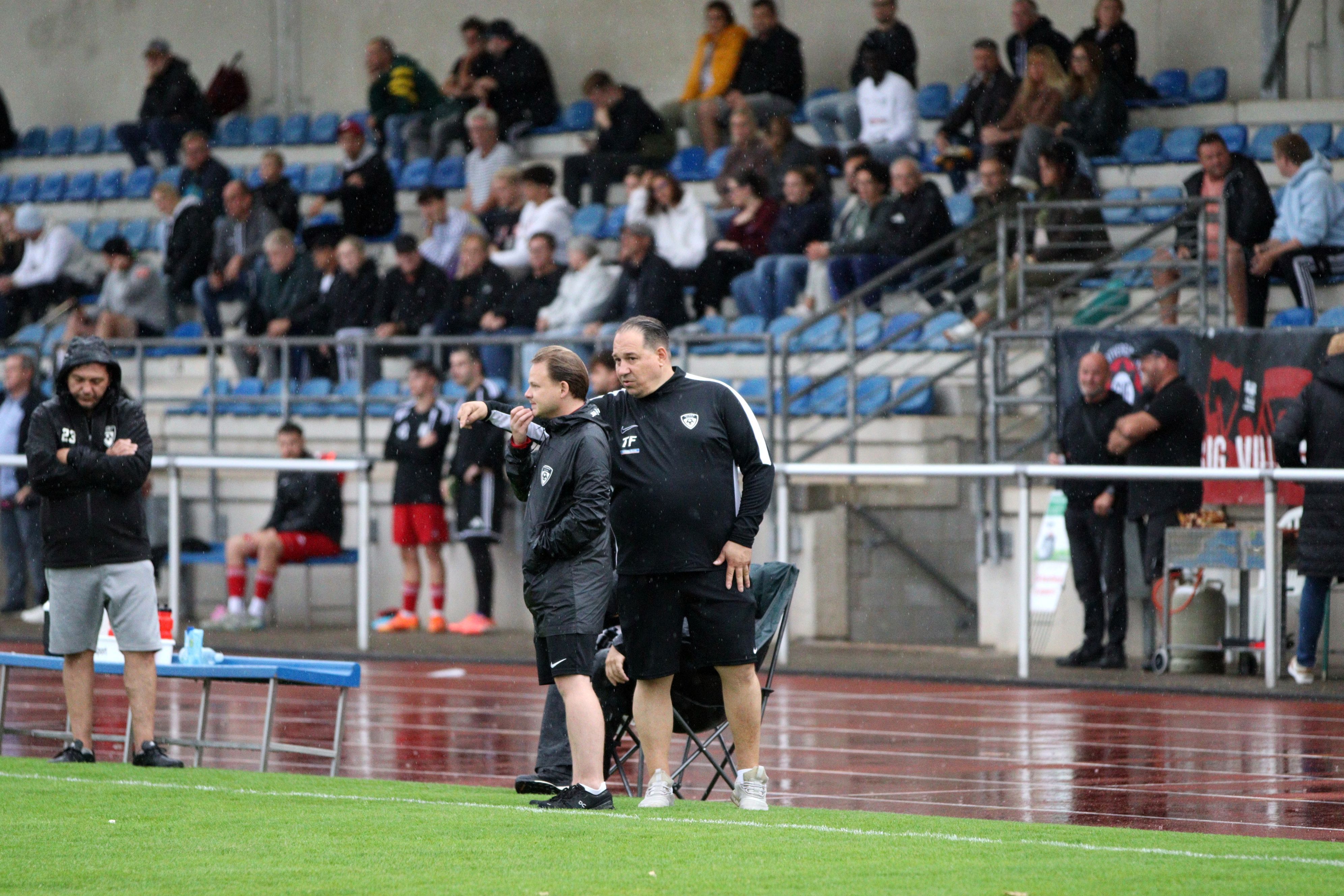 Erfts Co-Trainer spricht mit dem Cheftrainer und gestikuliert. Im Hintergrund die gut besetzte Tribüne des Heinz-Flohe-Stadions.