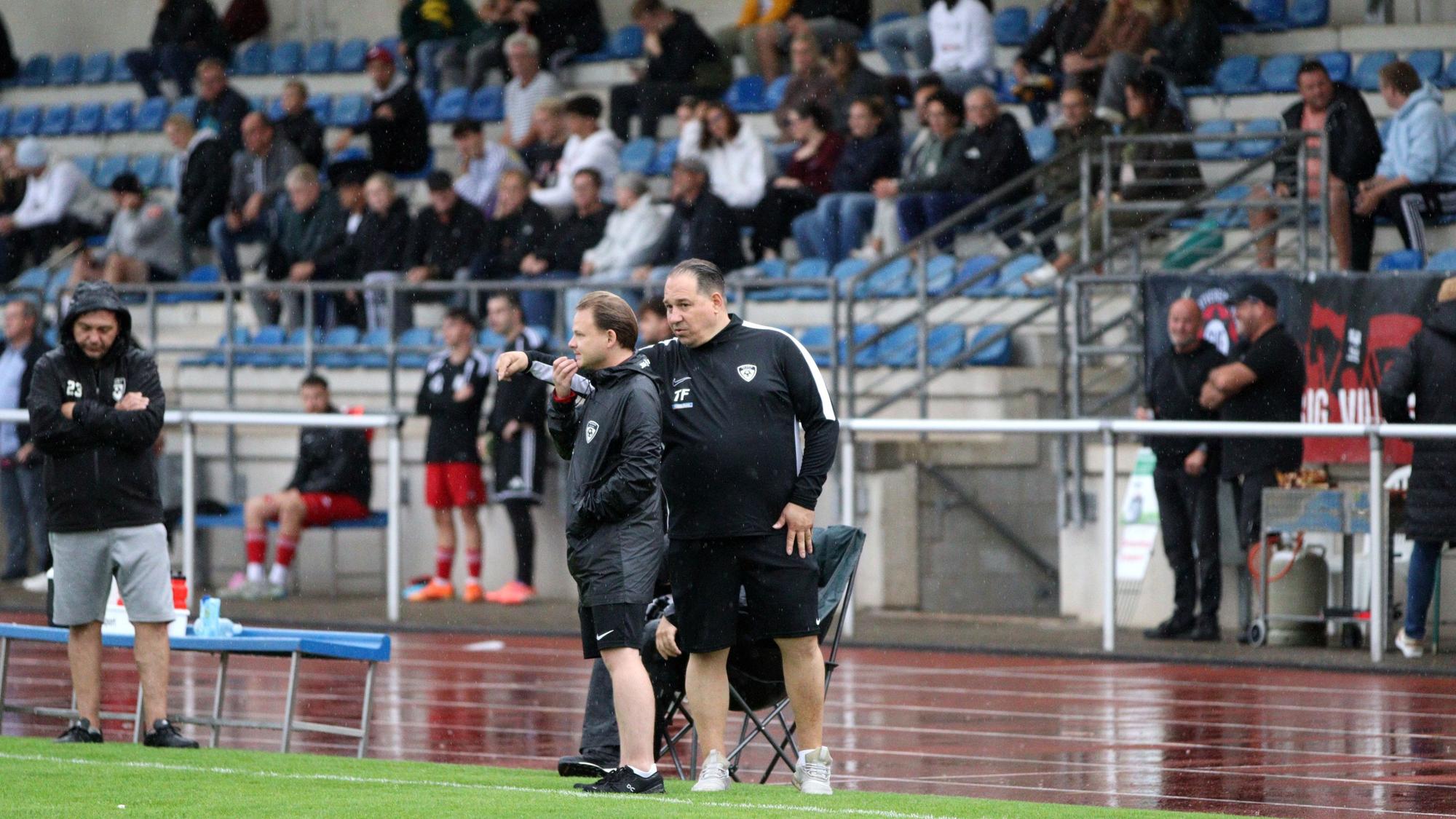Erfts Co-Trainer spricht mit dem Cheftrainer und gestikuliert. Im Hintergrund die gut besetzte Tribüne des Heinz-Flohe-Stadions.