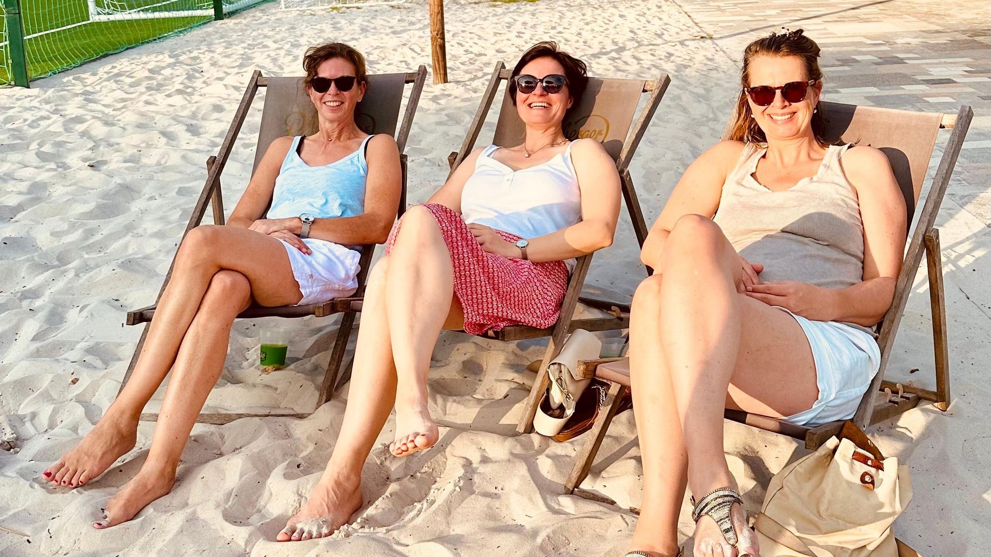 Zu sehen sind drei Frauen auf Strandstühlen auf einem Beachvolleyball-Feld.