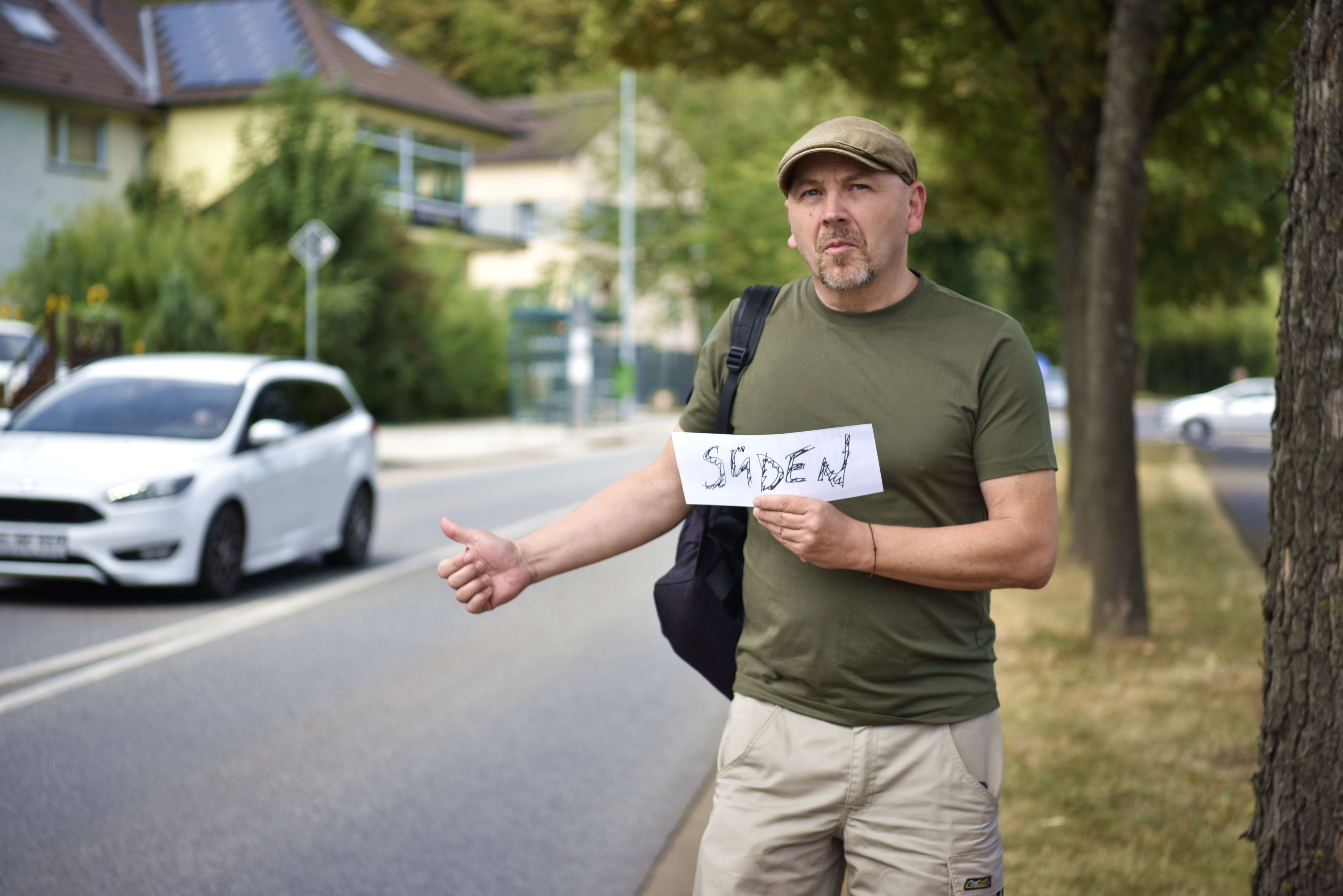 Thorsten Wirtz steht in grünem T-Shirt mit Rucksack und Schirmmütze an einer Straße und hält einen Daumen nach oben. In der anderen Hand hält er ein Schild auf dem "Süden" steht.