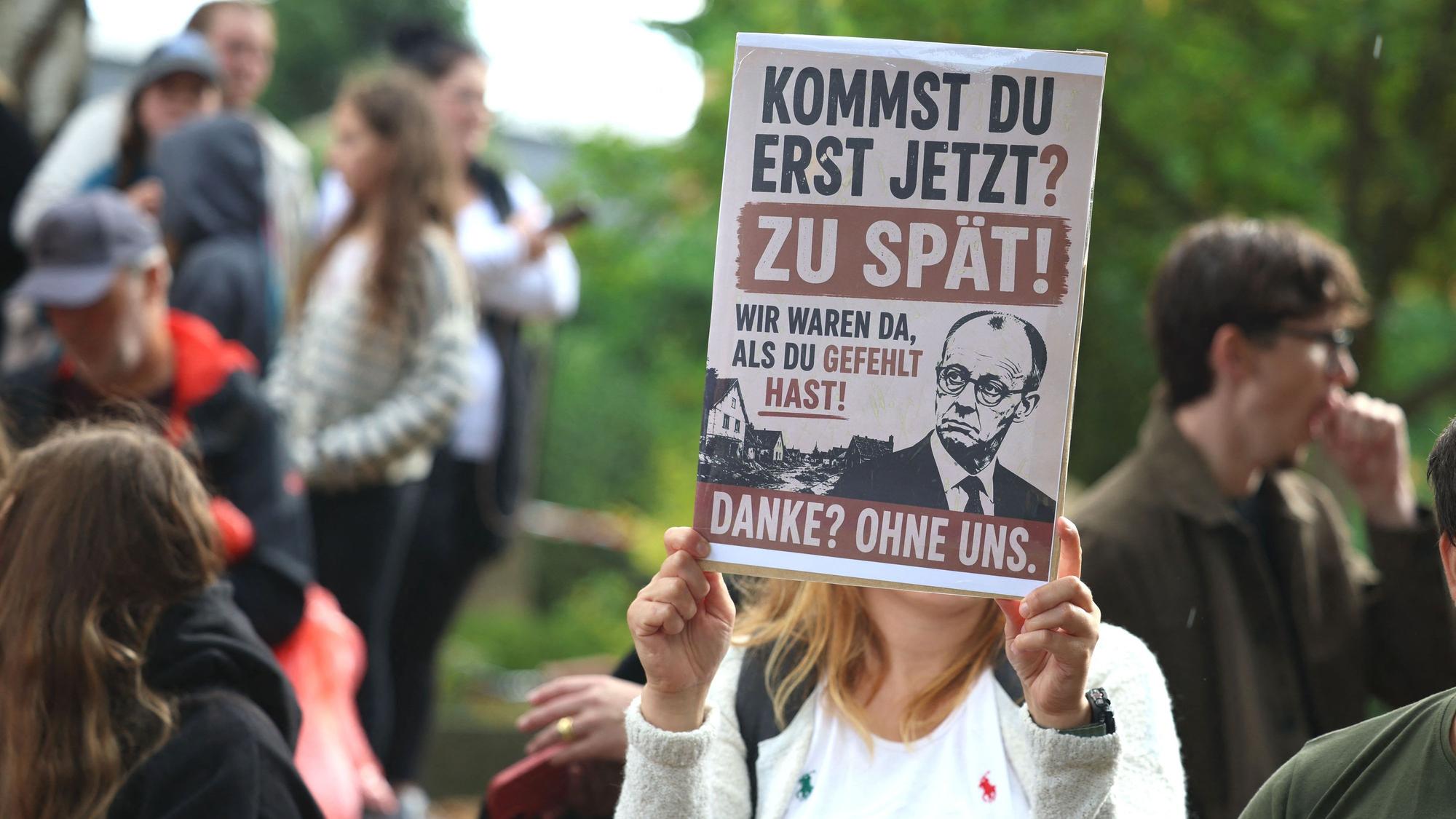 Eine Frau hält ein Plakat mit der Aufschrift „Kommst du erst jetzt? Zu spät!“ bei einem Besuch von Bundeskanzler Merz in Hürtgenwald-Gey.