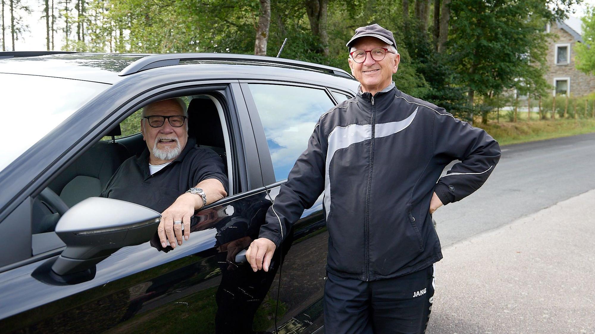 Das Foto zeigt zwei ältere Männer: Einer sitzt bei geöffnetem Fenster am Steuer eines schwarzen Autos, der andere steht daneben und lehnt sich mit einem Arm an das Fahrzeug.