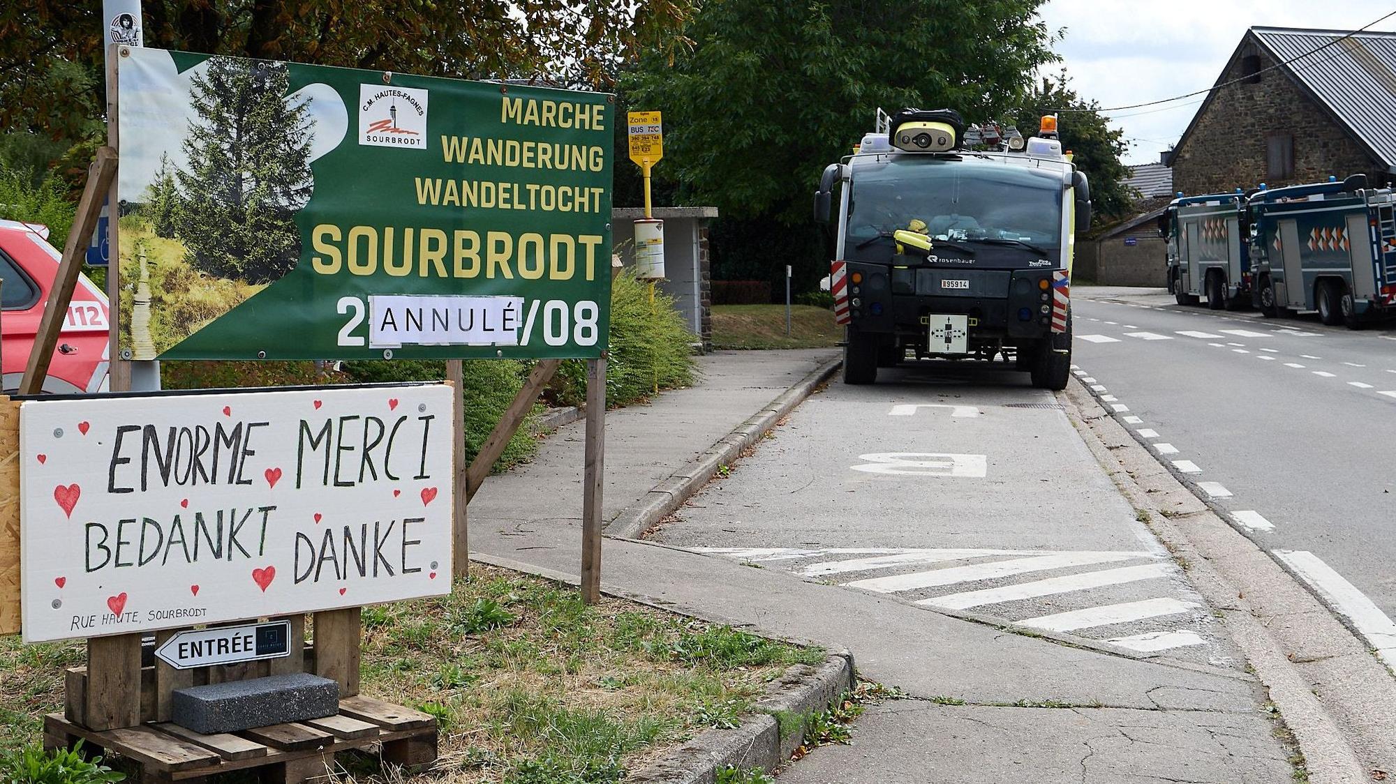 In Sourbrodt parken zwei große Einsatzfahrzeuge am Straßenrand, im Vordergrund sind Schilder zu sehen, die auf eine abgesagte Veranstaltung hinweisen und in mehreren Sprachen den Einsatzkräften gedankt wird.