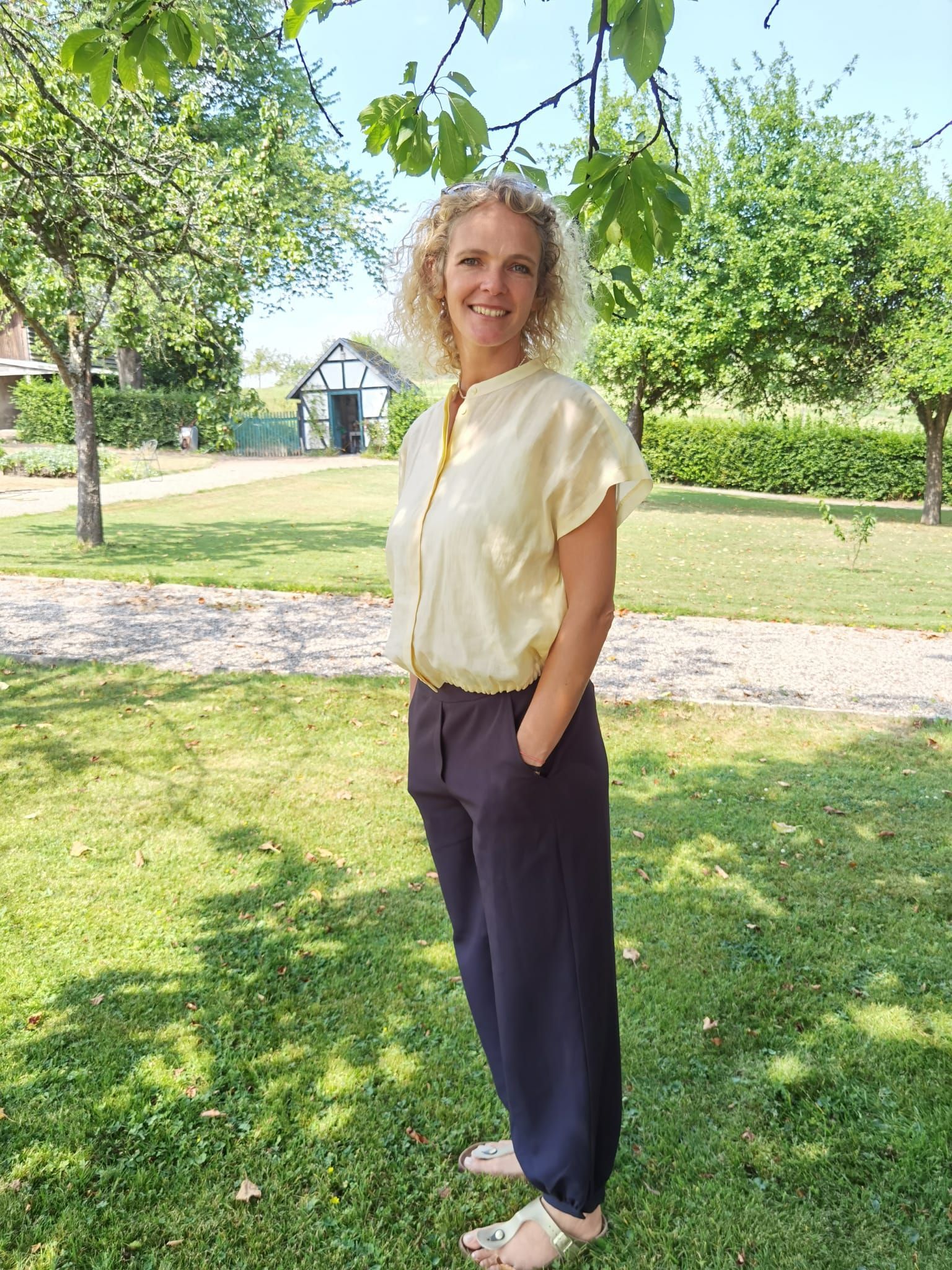 Eine Frau mit blonden Locken steht in einem gepflegten Garten und lächelt.