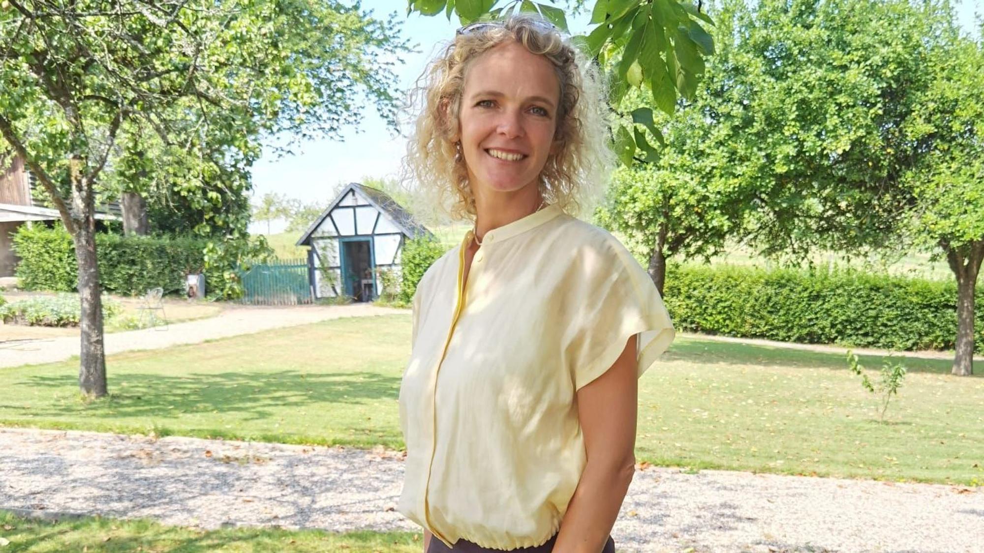 Eine Frau mit blonden Locken steht in einem gepflegten Garten und lächelt.