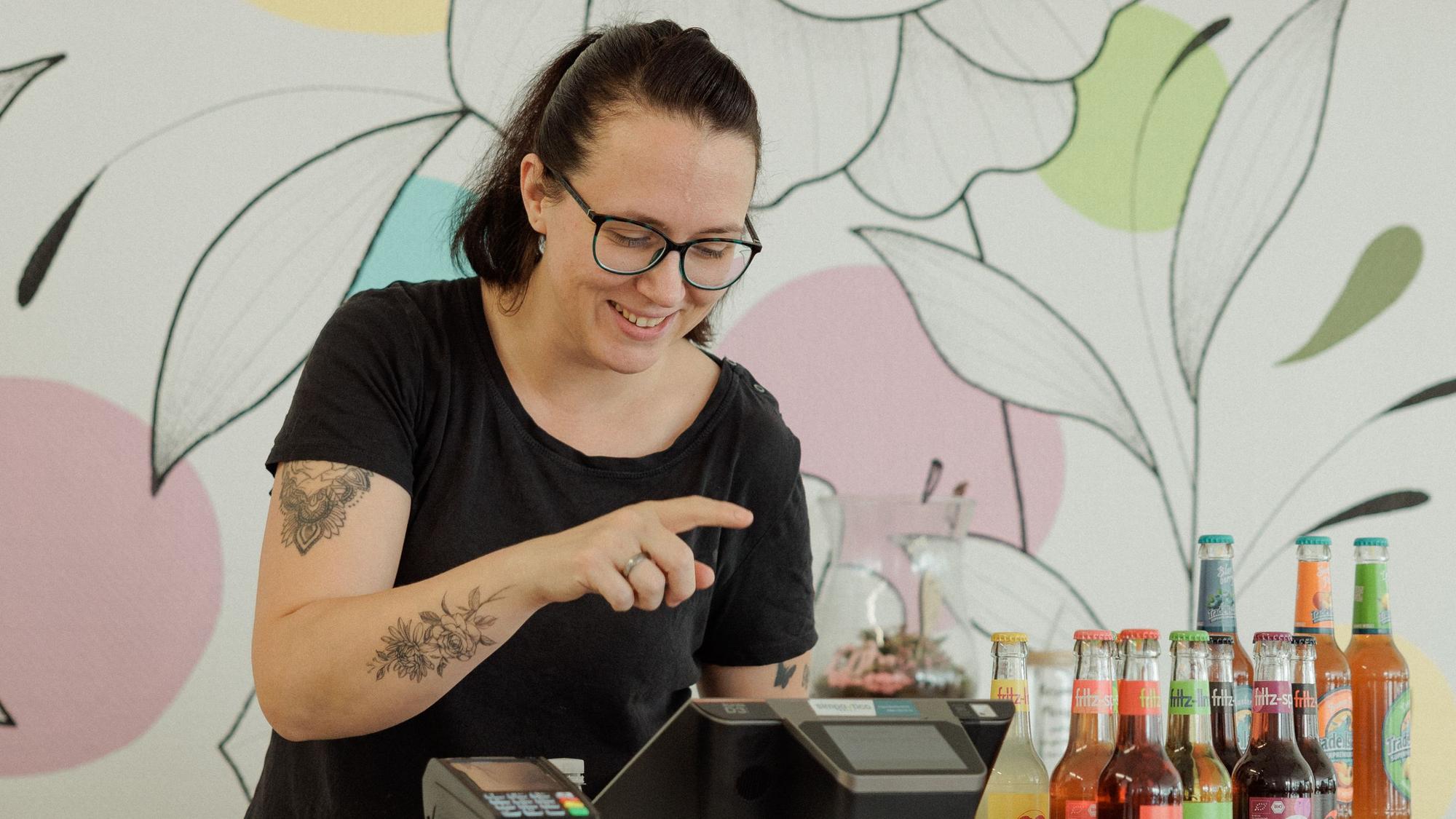 Zu sehen ist eine Frau, die hinter einem Café-Tresen steht und etwas in das Kassensystem eingibt.