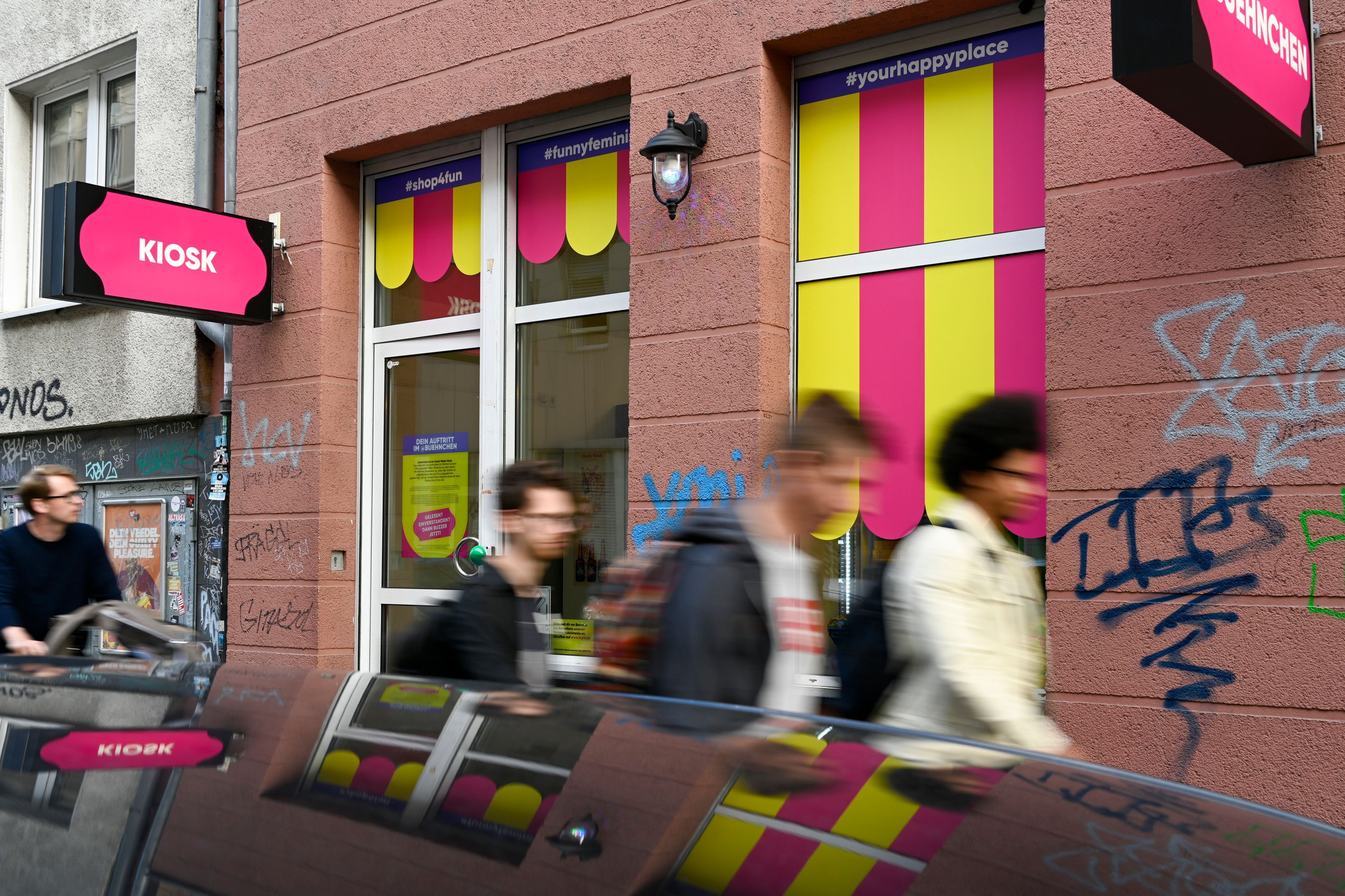 Der neue Kiosk in der Kyffhäuserstraße 15 hat am vergangenen Wochenende seine Eröffnung gefeiert. Die Schilder sind pink, es laufen mehrere Menschen daran vorbei.