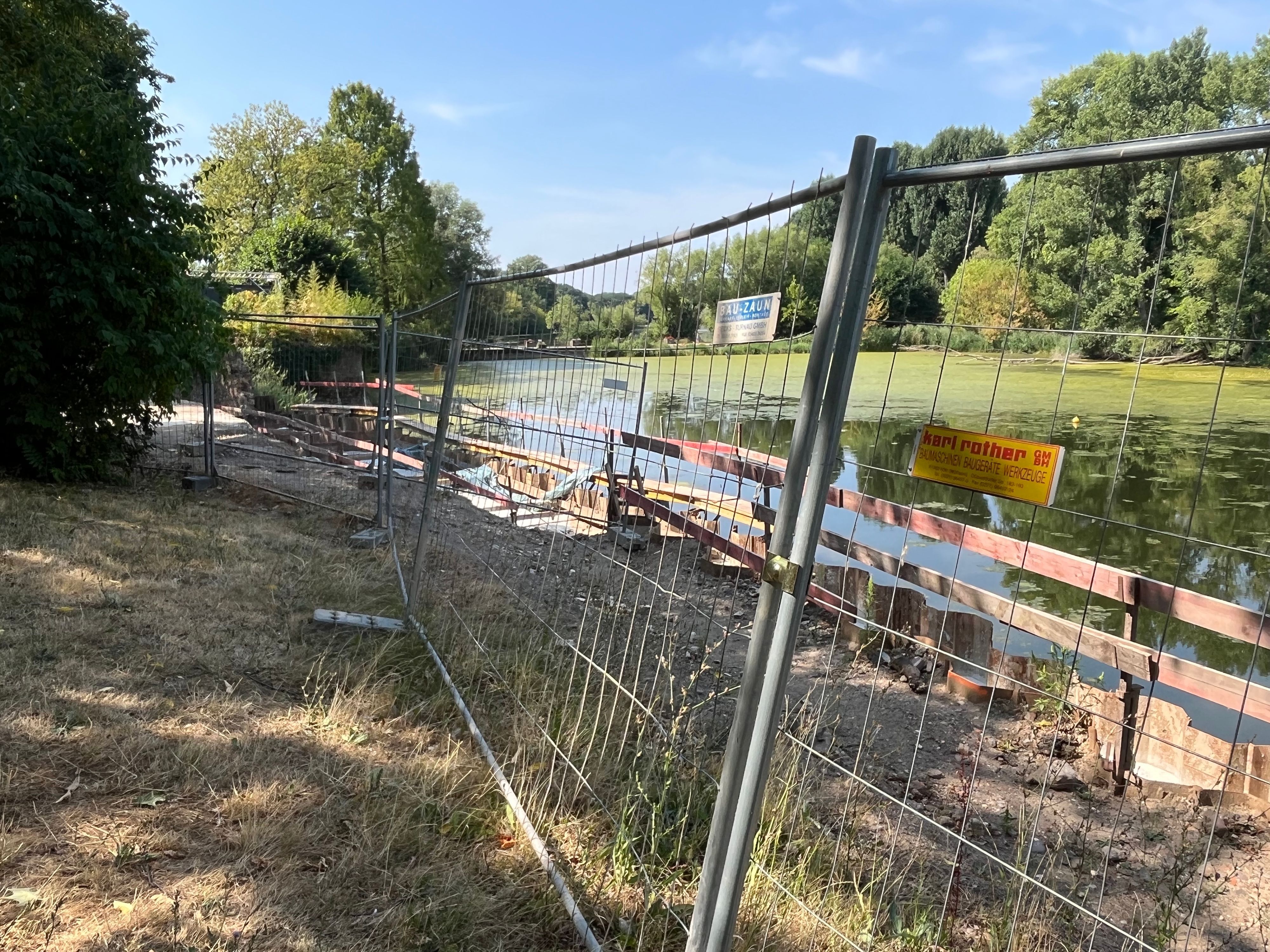 Hinter einem Bauzaun ist ein Teich zu sehen.