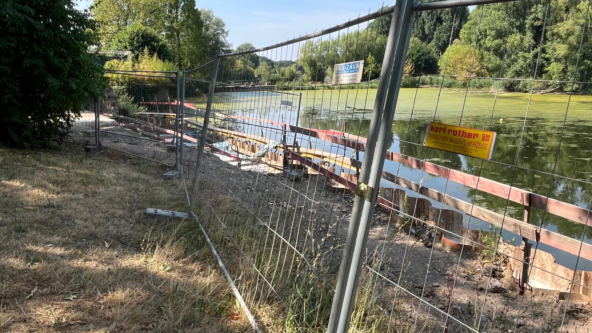 Hinter einem Bauzaun ist ein Teich zu sehen.