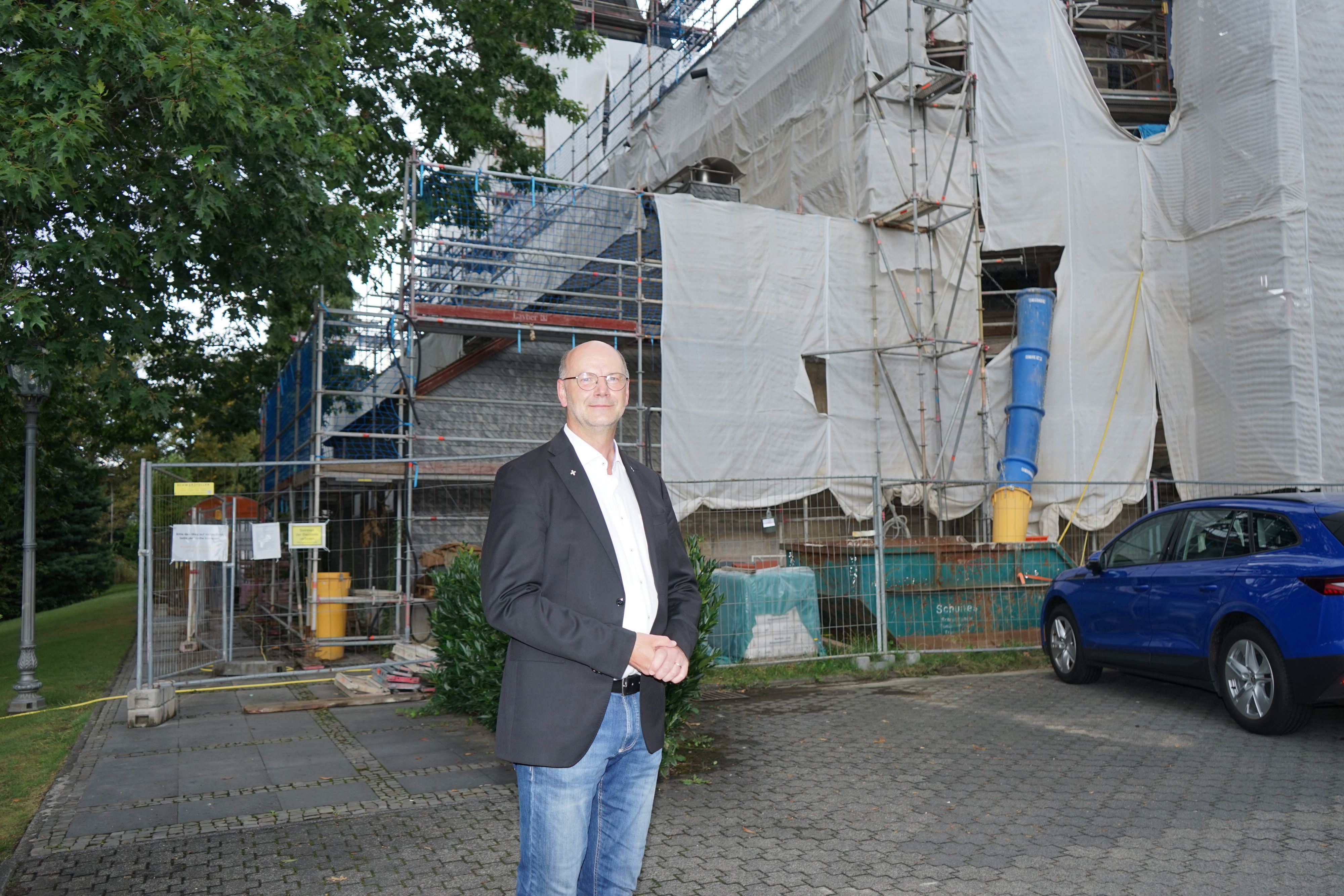 ein Mann in weißem Hemd und dunklem Sakko steht vor einem eingerüsteten Gebäude