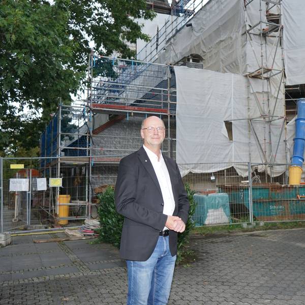 ein Mann in weißem Hemd und dunklem Sakko steht vor einem eingerüsteten Gebäude