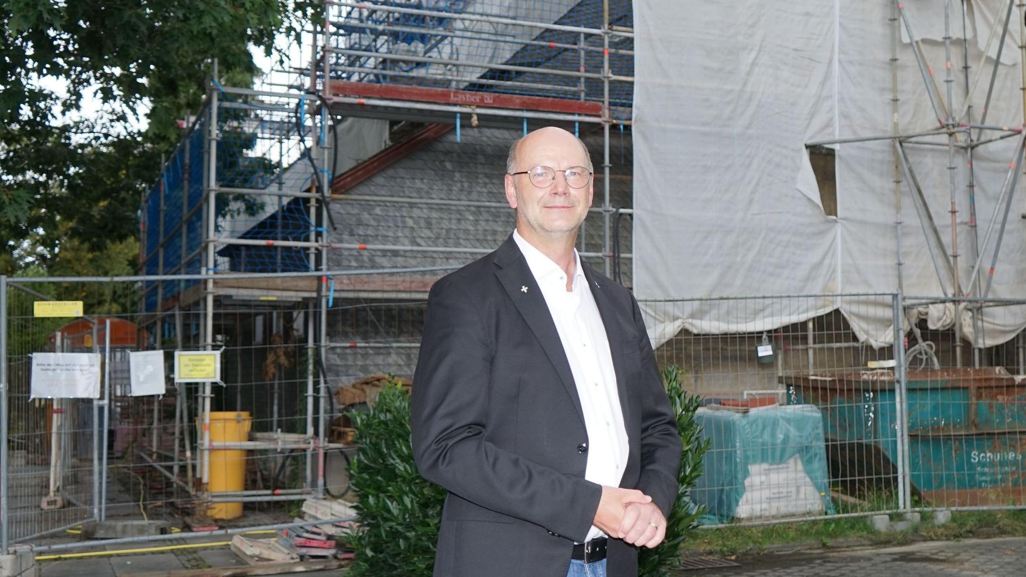 ein Mann in weißem Hemd und dunklem Sakko steht vor einem eingerüsteten Gebäude