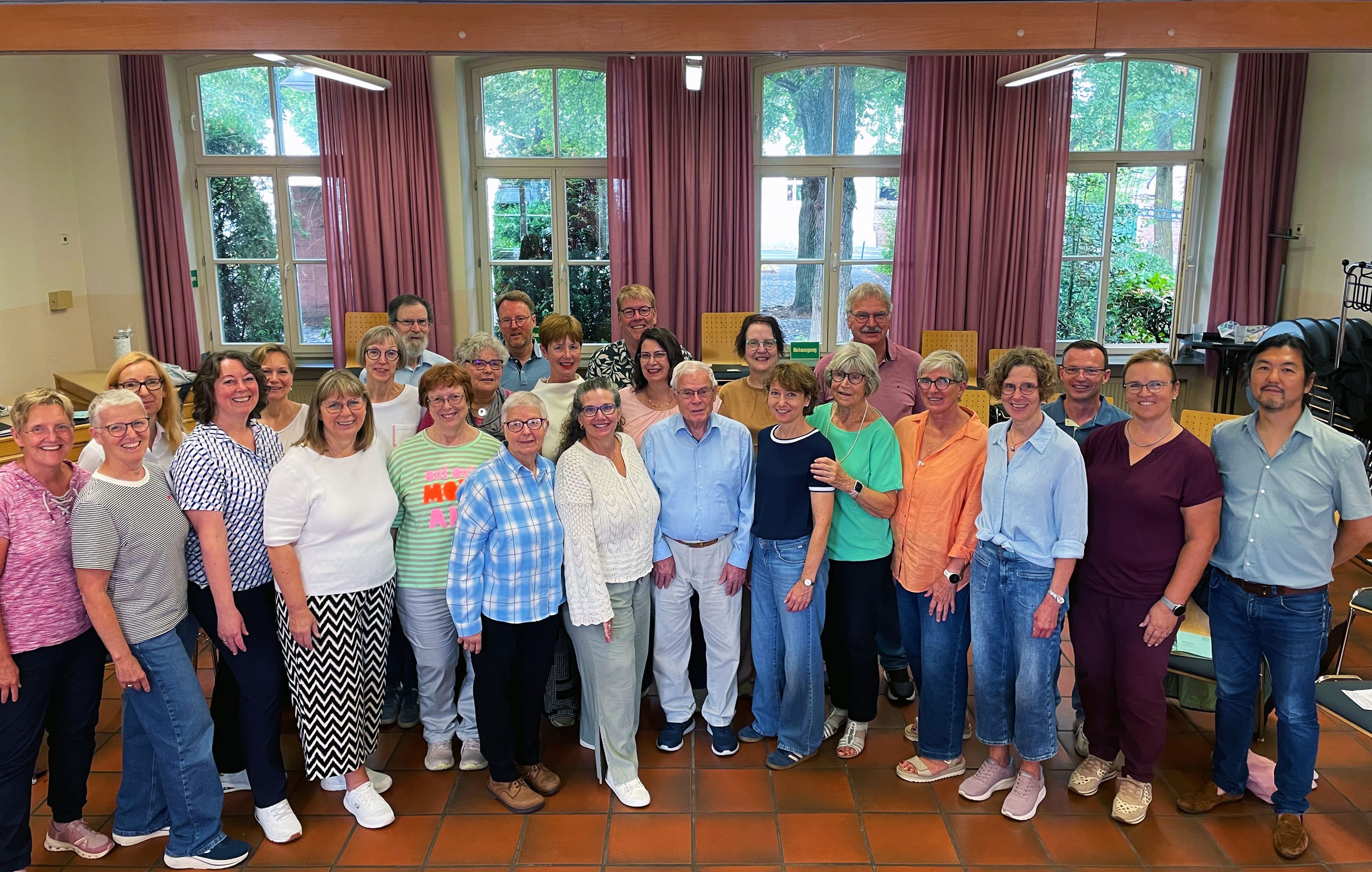 Eine bunt gemischte Gruppe von Männern und Frauen steht im Halbkreis in einem großen Raum vor geöffneten Fenstern.