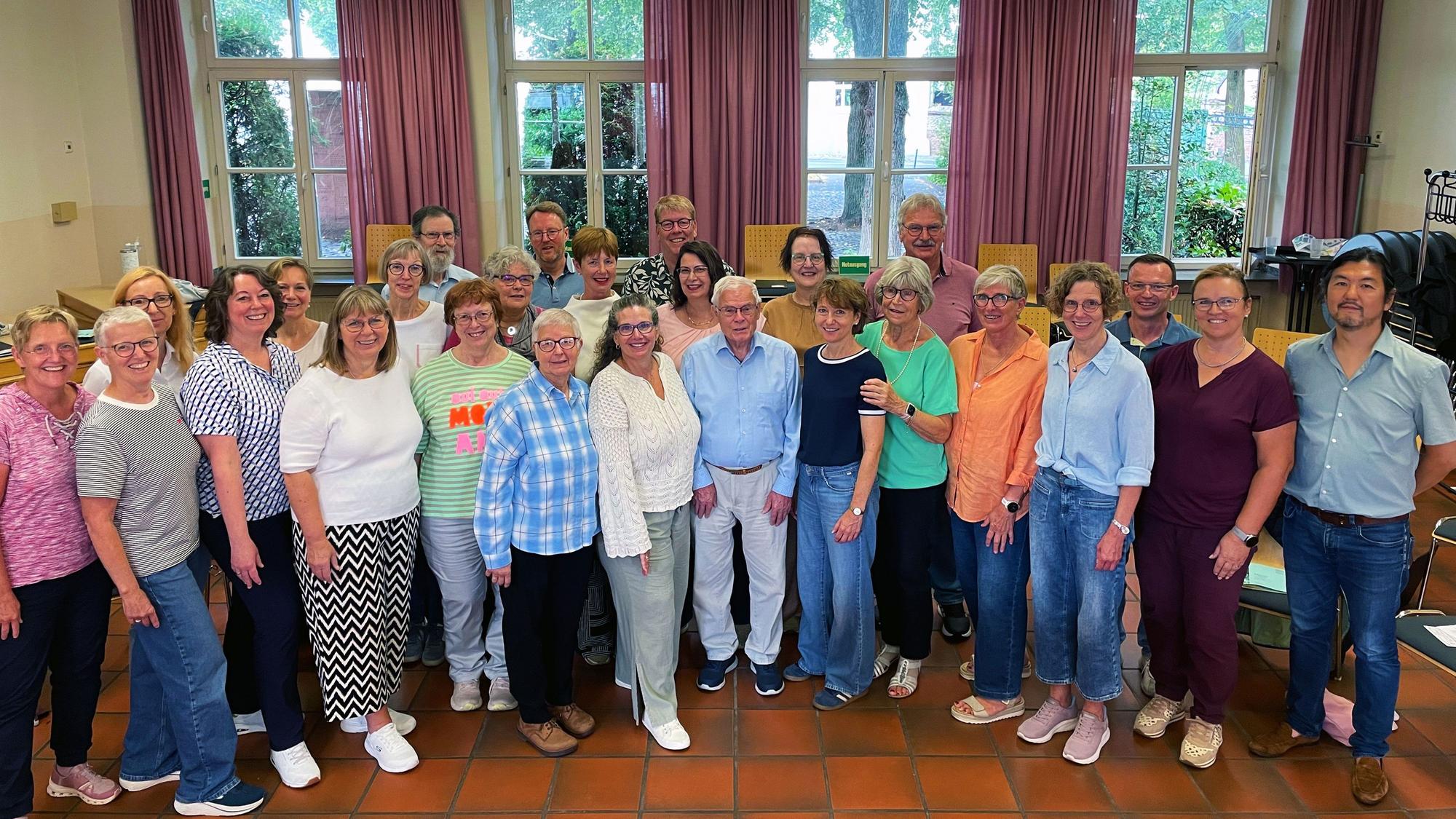 Eine bunt gemischte Gruppe von Männern und Frauen steht im Halbkreis in einem großen Raum vor geöffneten Fenstern.