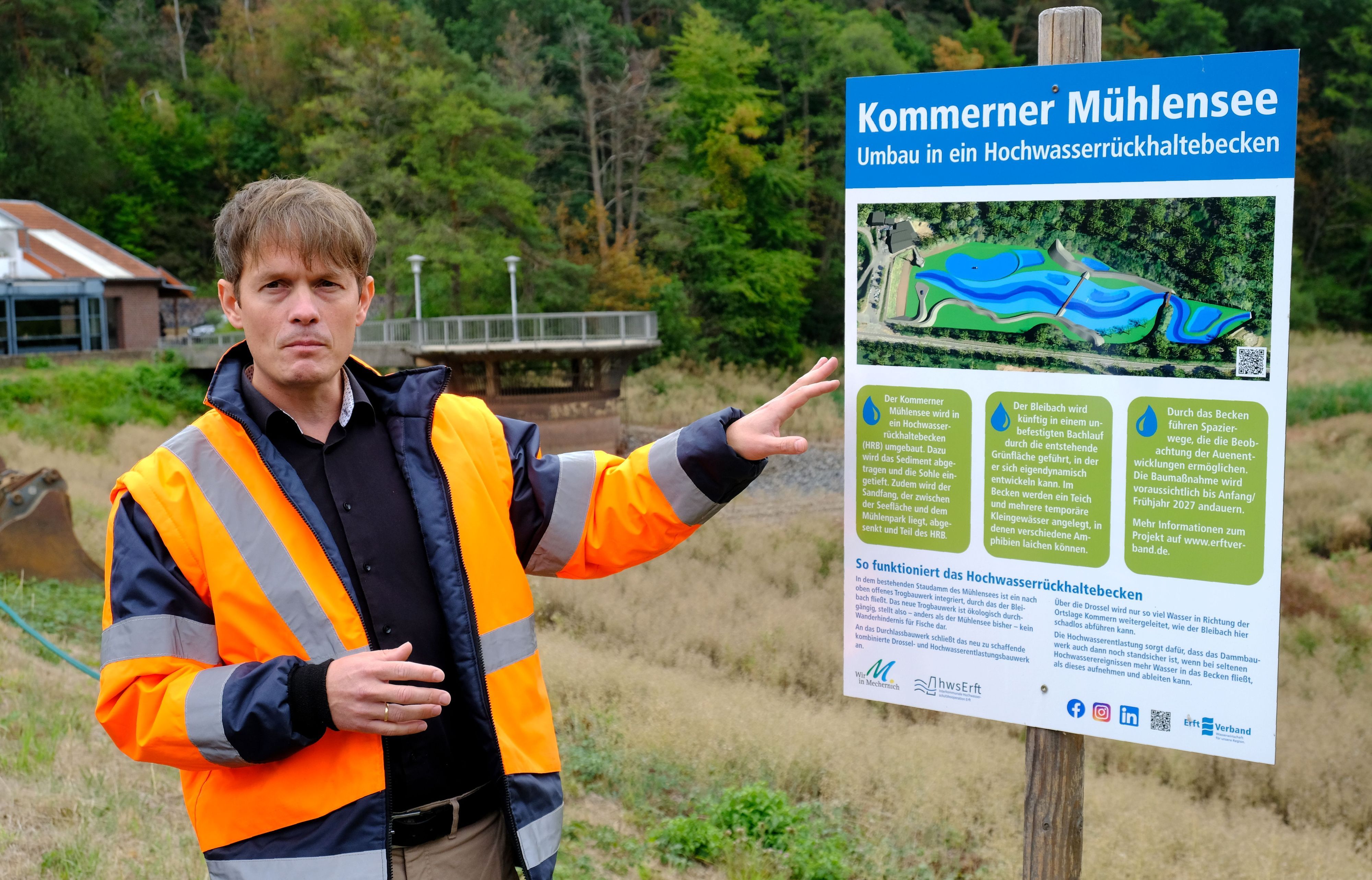 Ein Mann, der eine orangefarbene Signaljacke trägt, deutet auf eine Schautafel mit der Aufschrift „Kommerner Mühlensee - Umbau in ein Hochwasserrückhaltebecken“.