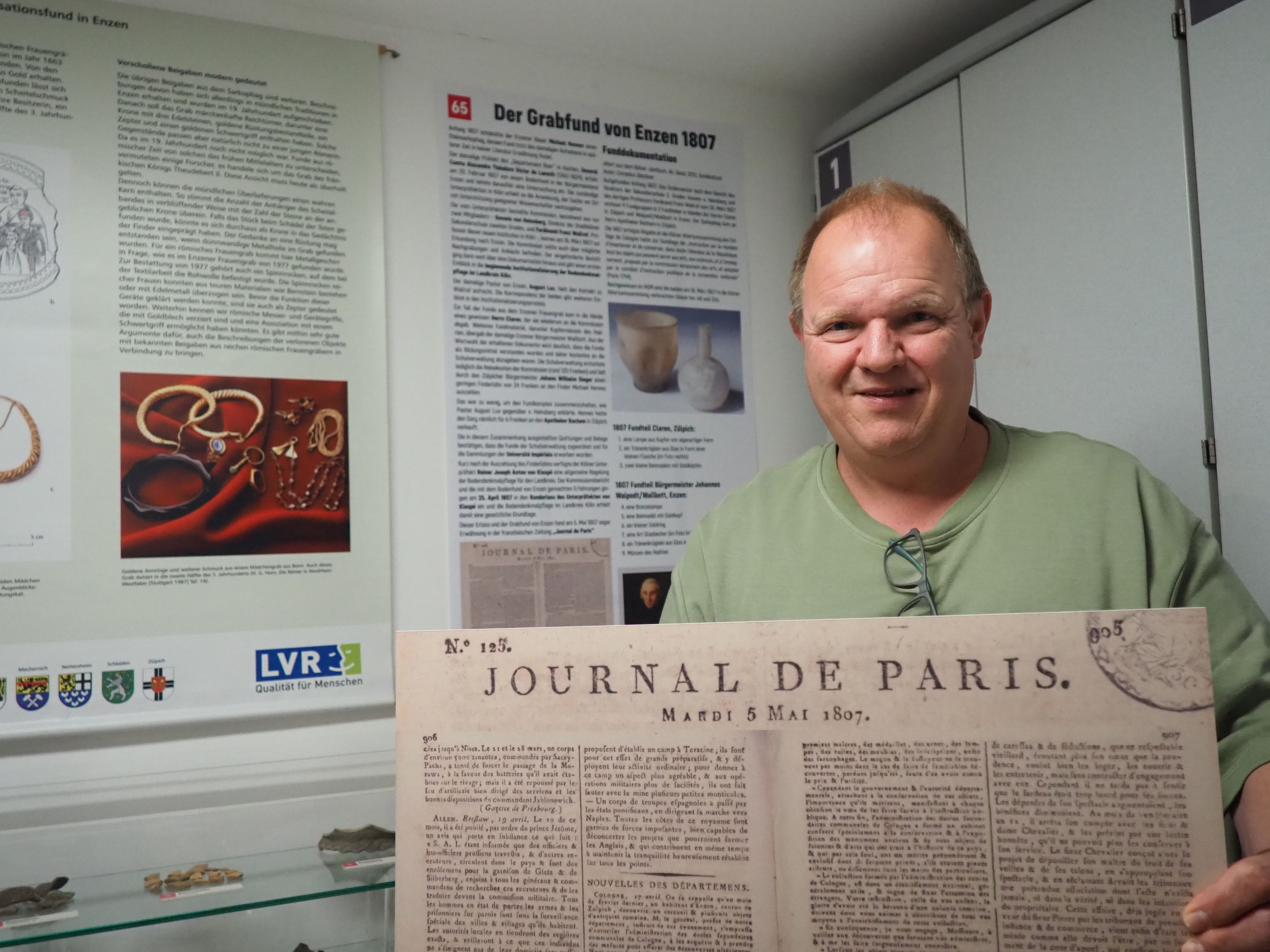 Ein Mann steht vor Schautafeln und präsentiert eine historische Zeitungsseite des „Journal de Paris“.