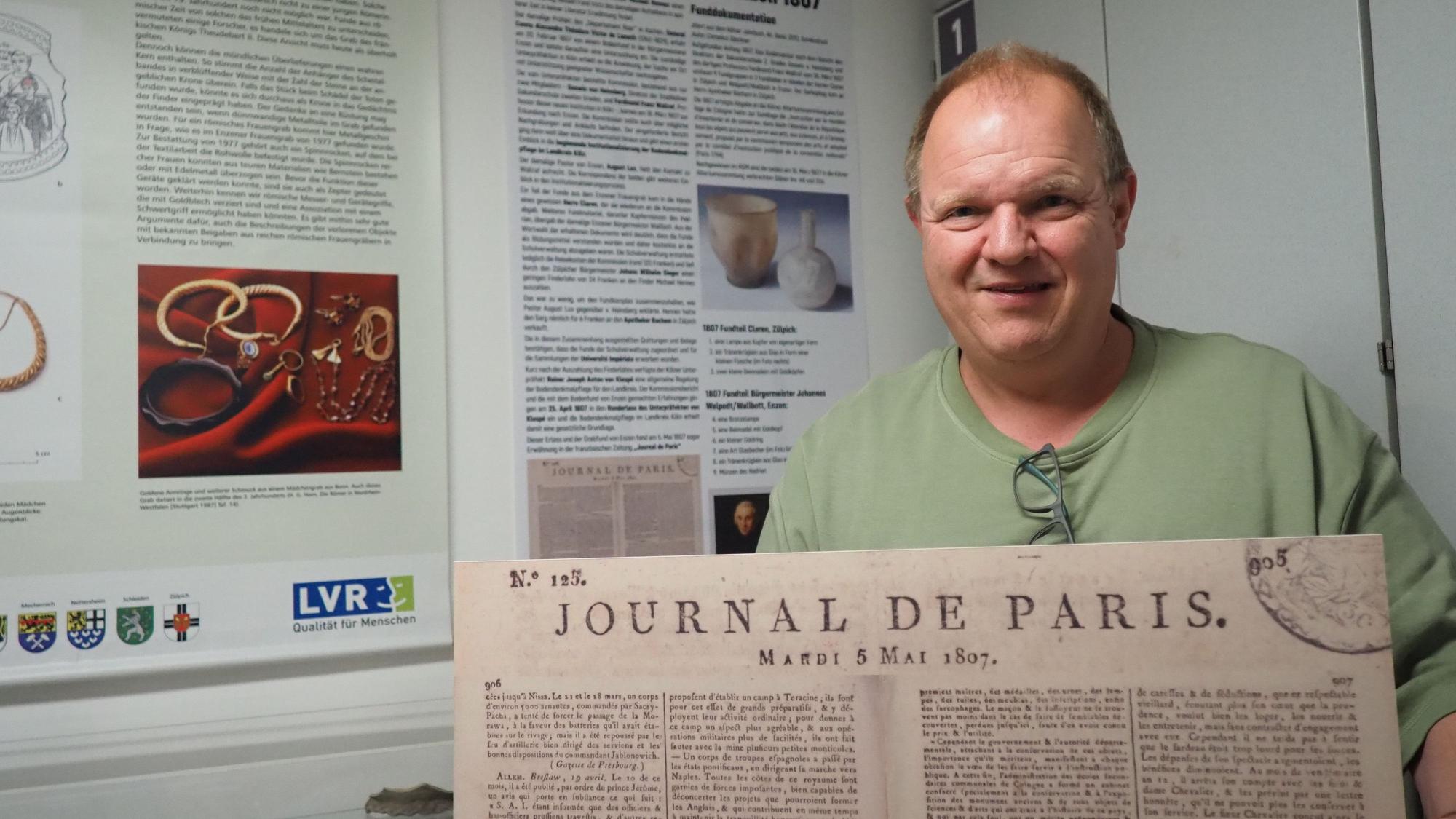 Ein Mann steht vor Schautafeln und präsentiert eine historische Zeitungsseite des „Journal de Paris“.