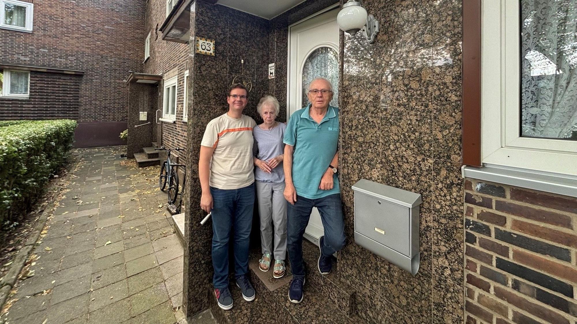 Auf dem Bild sind drei Personen vor einem Haus zu sehen.
