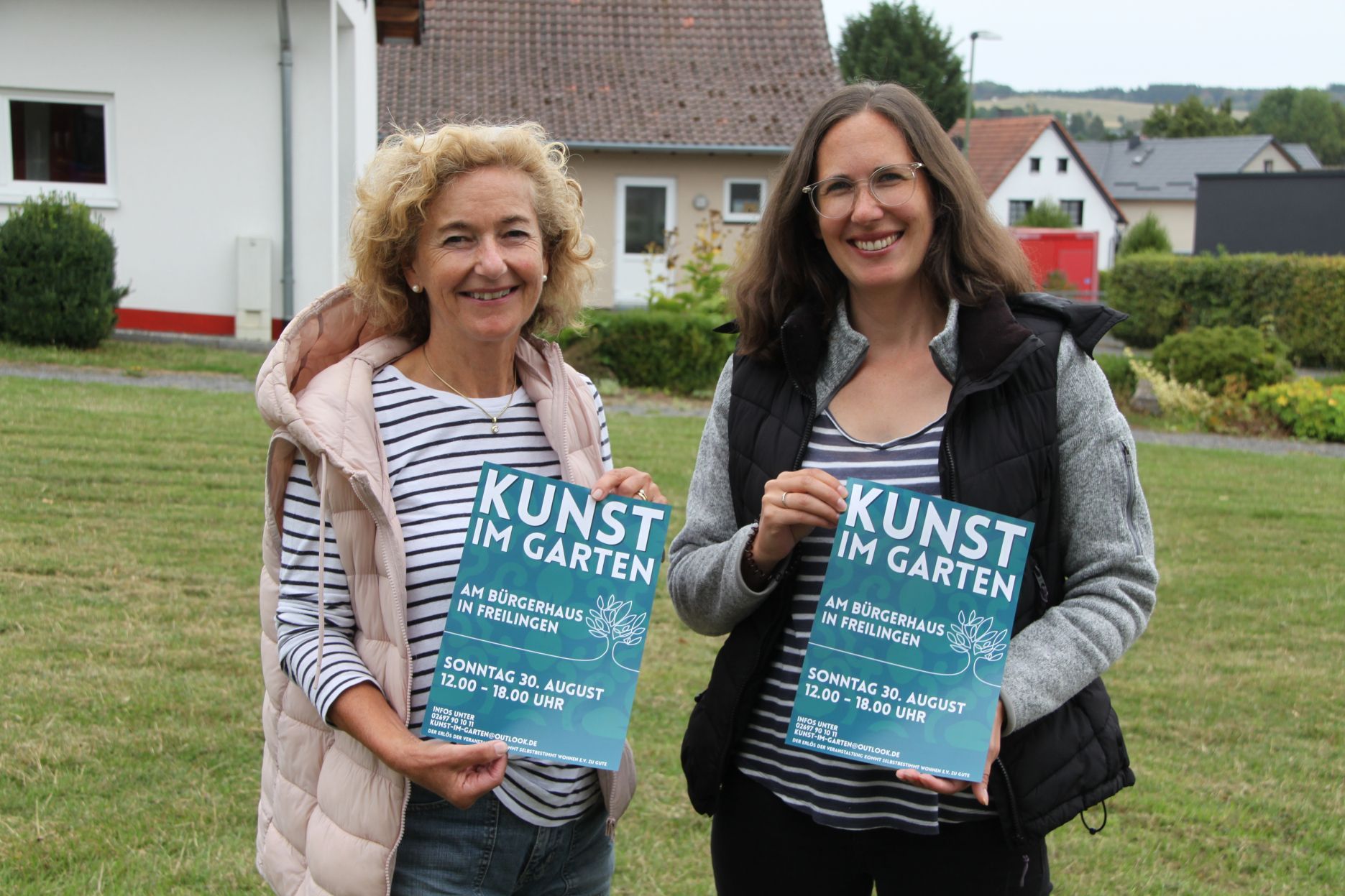 Die beiden genannten Frauen stehen nebeneinander und präsentieren jeweils ein Veranstaltungsplakat von „Kunst im Garten“.