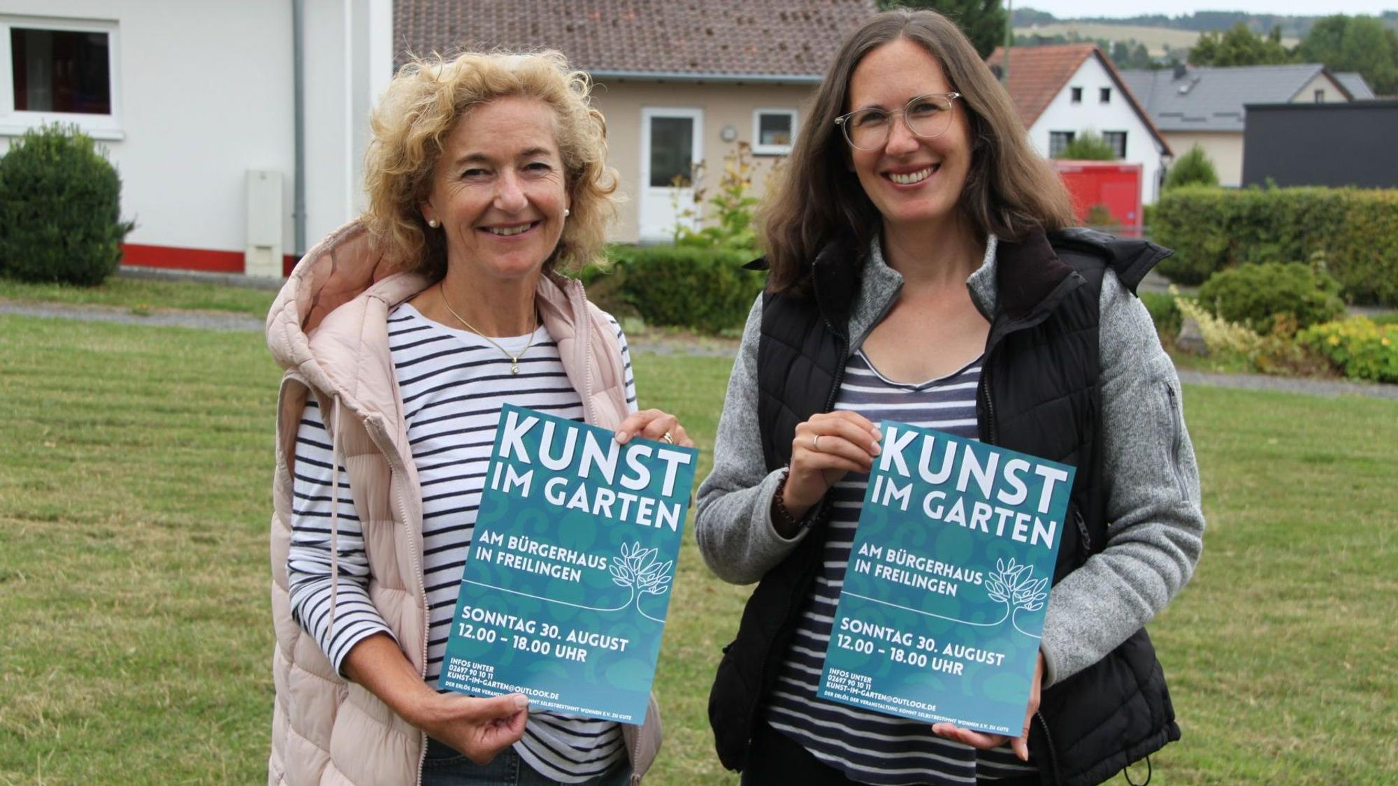 Die beiden genannten Frauen stehen nebeneinander und präsentieren jeweils ein Veranstaltungsplakat von „Kunst im Garten“.