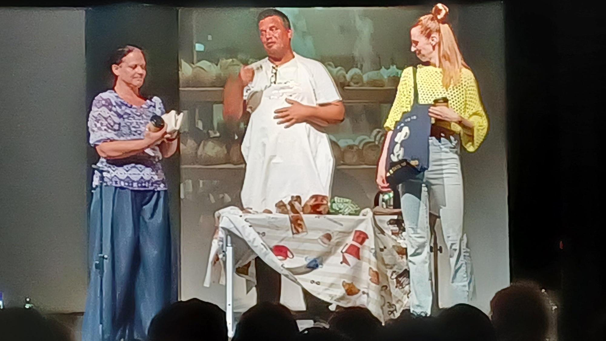 Zwei Frauen und ein Mann stehen auf einer Bühne, zwischen sich ein Tisch, im Hintergrund sind Brote im Regal zu sehen.
