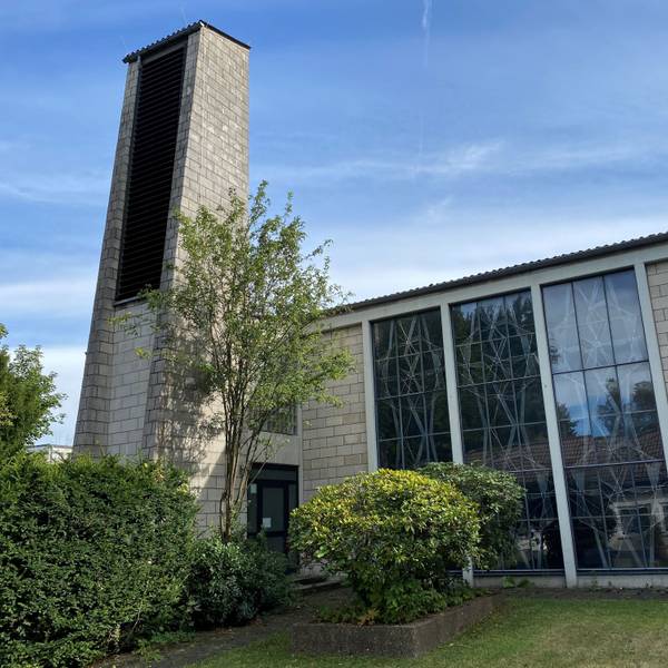 Ein weißes Kirchengebäude mit angebautem Glockenturm aus den 1970er Jahren, davor grüne Sträucher.