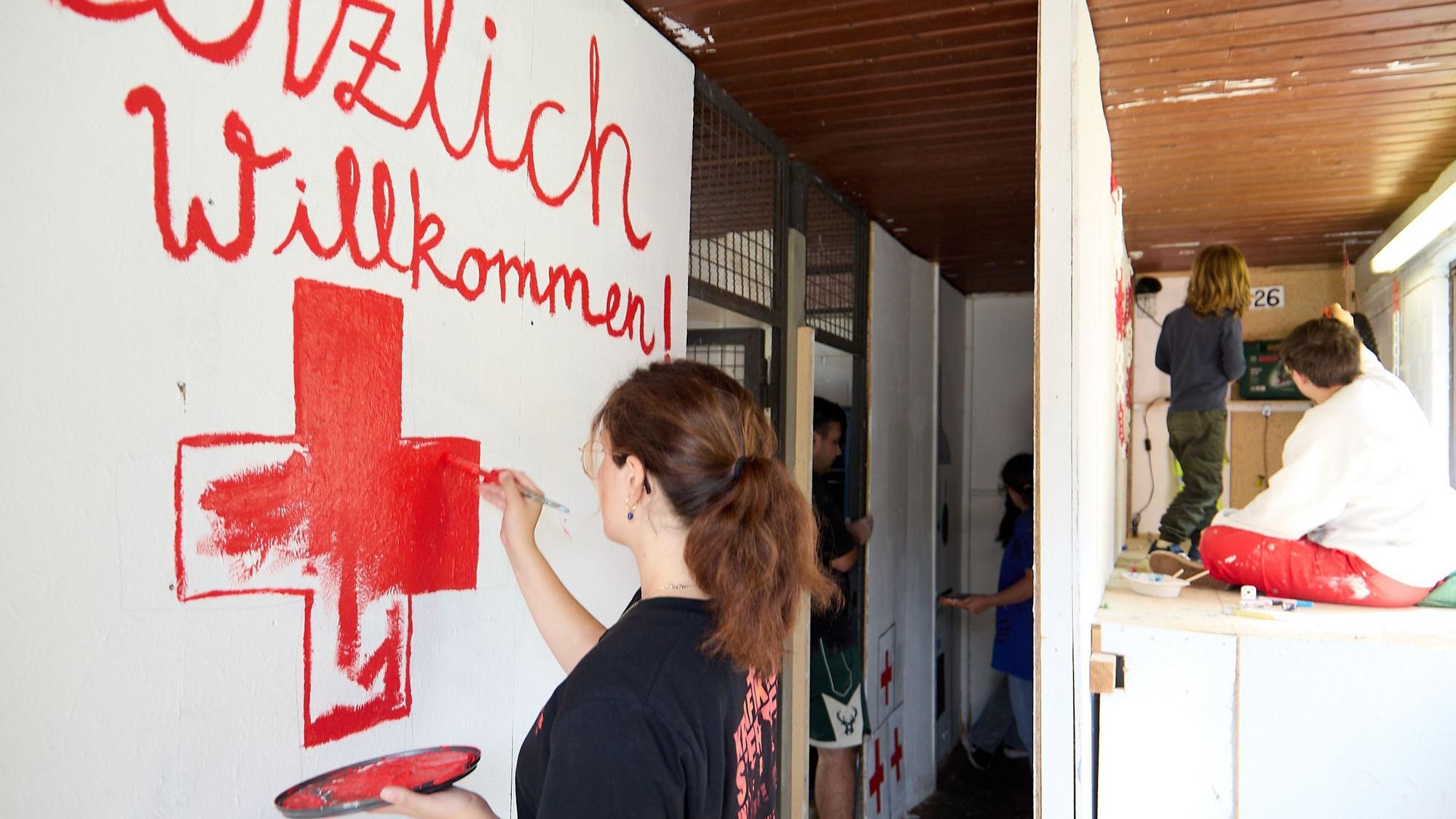 Eine junge Frau pinselt mit roter Frabe ein Kreuz auf eine weiße Wand. Darüber ist der Schriftzug „Herzlich Willkommen“ zu sehen.