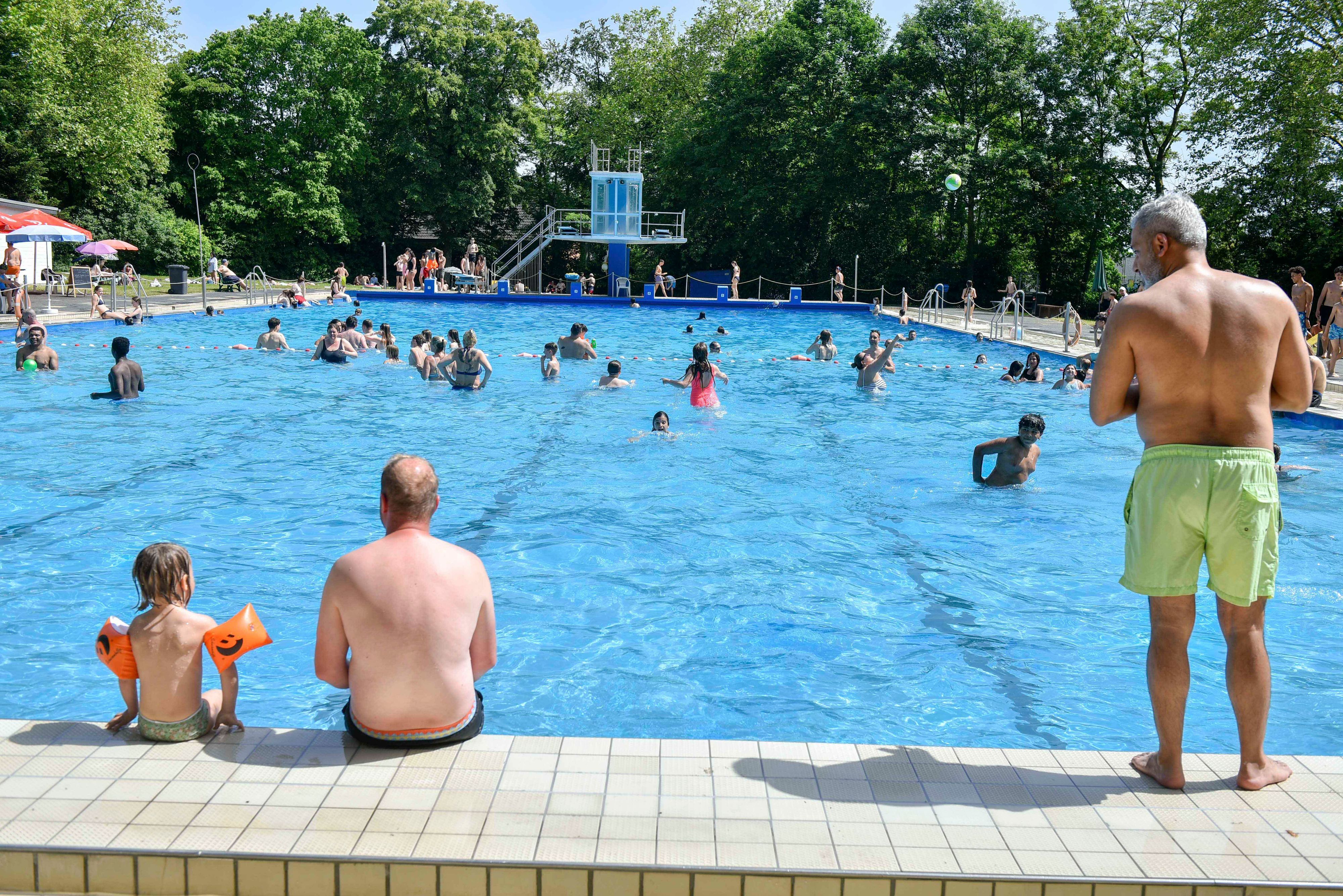 Menschen sitzen am Beckenrand eines Freibads, viele weitere plantschen im Wasser.