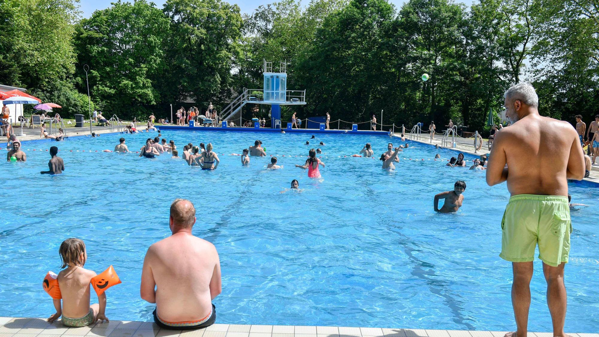 Menschen sitzen am Beckenrand eines Freibads, viele weitere plantschen im Wasser.