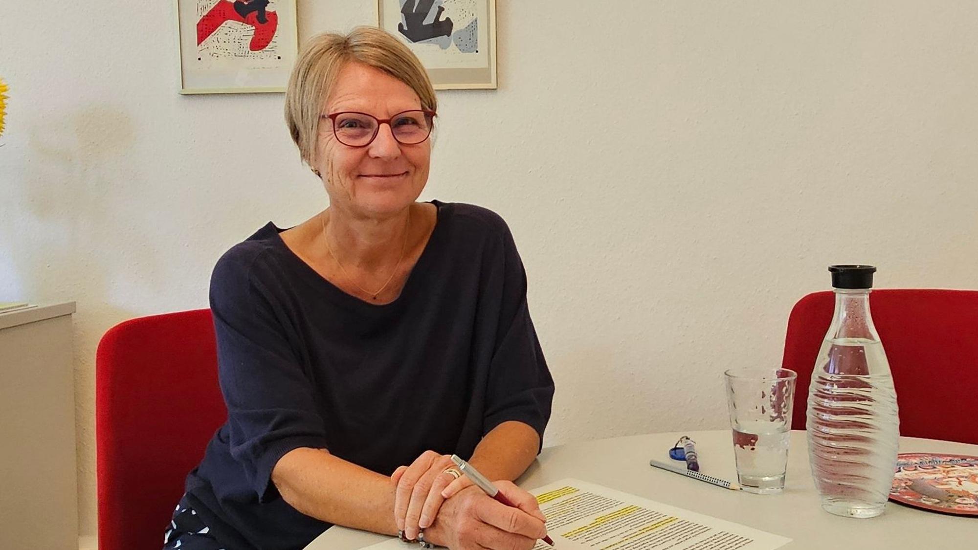 Auf dem Bild ist das Portrait einer Frau zu sehen, sie sitzt an einem Tisch.