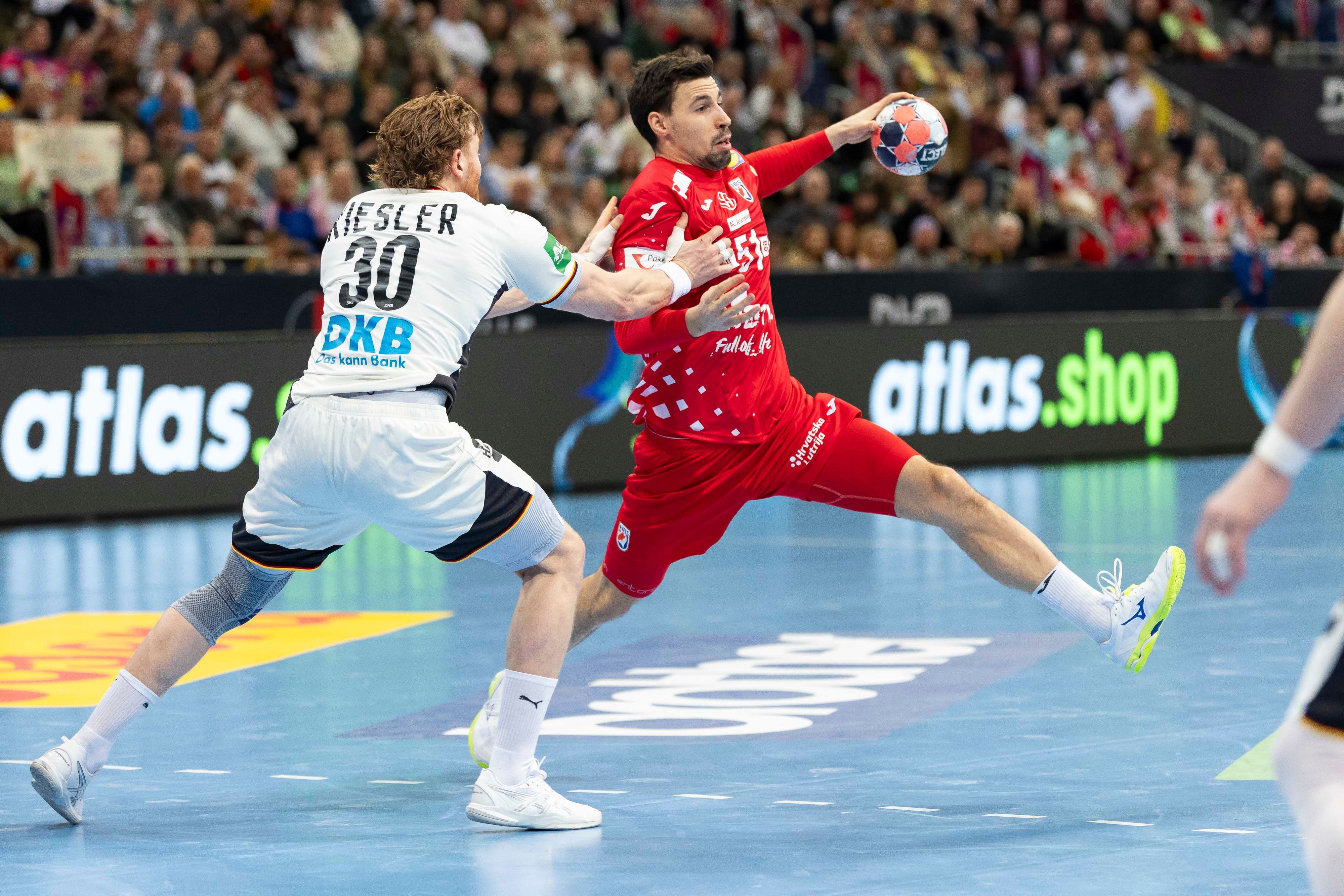 Ein Handballspieler im roten Trikot versucht, sich mit dem Ball gegen einen Gegenspieler im weißen Trikot durchzusetzen. Im Hintergrund ist eine jubelnde Zuschauermenge zu sehen.