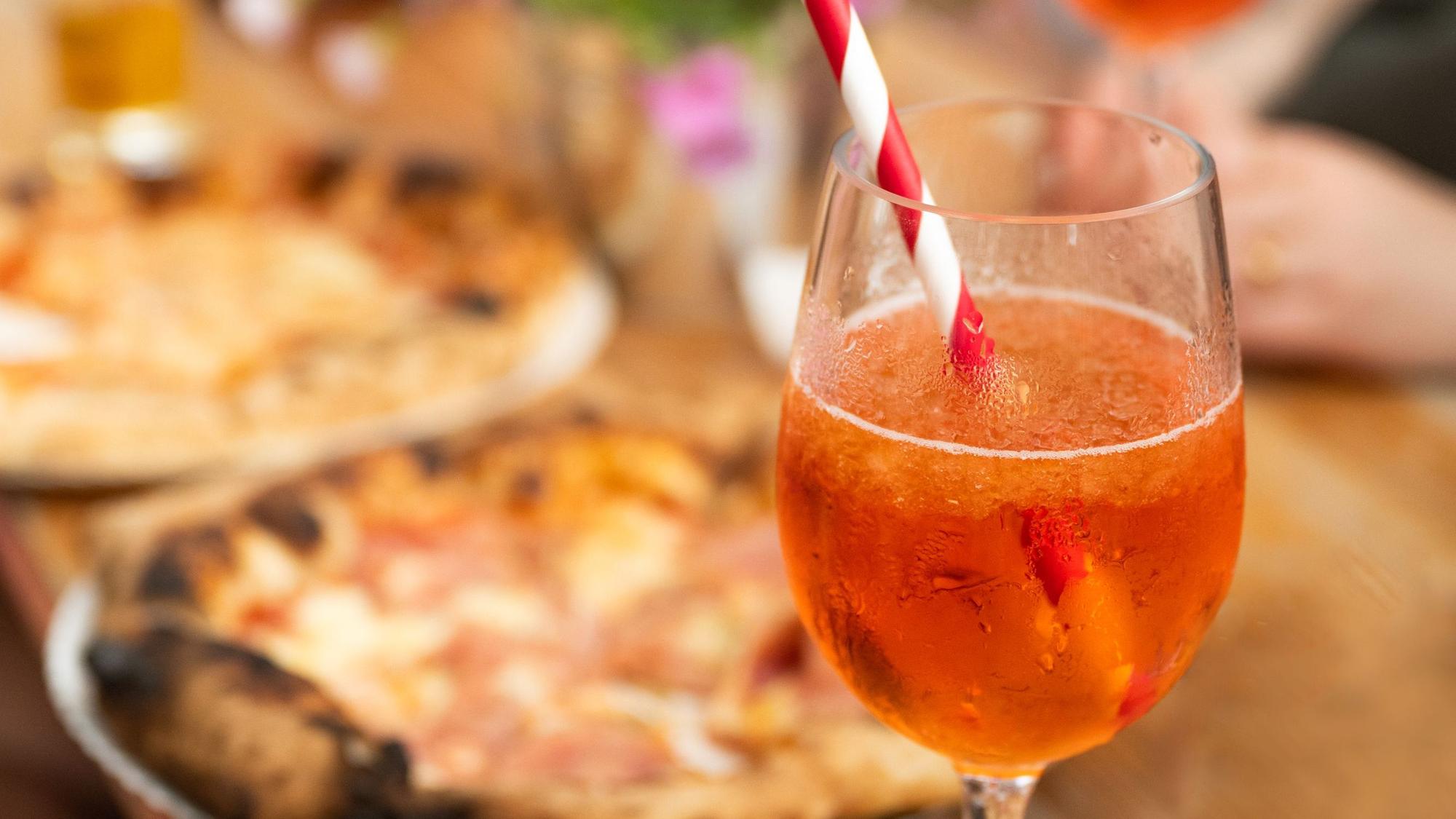 Ein Glas Aperol Spritz steht vor zwei Pizzen auf einem hölzernen Tisch. (Symbolbild)