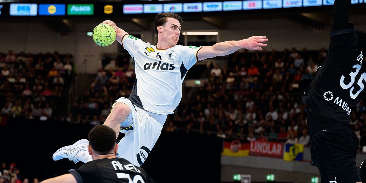 Ein deutscher Handballspieler im weißen Trikot springt zum Wurf, während zwei Gegenspieler ihn zu blocken versuchen.