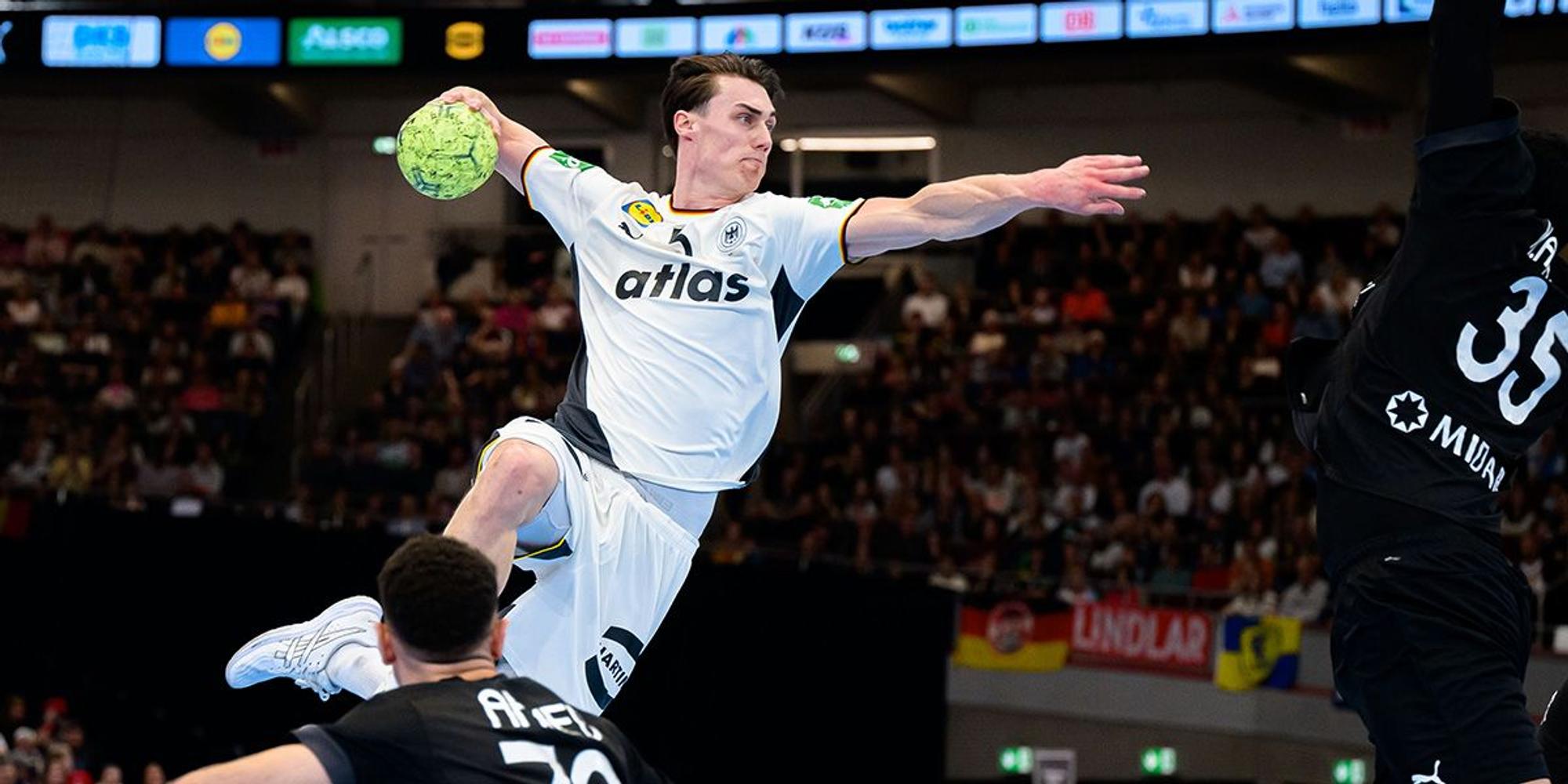 Ein deutscher Handballspieler im weißen Trikot springt zum Wurf, während zwei Gegenspieler ihn zu blocken versuchen.