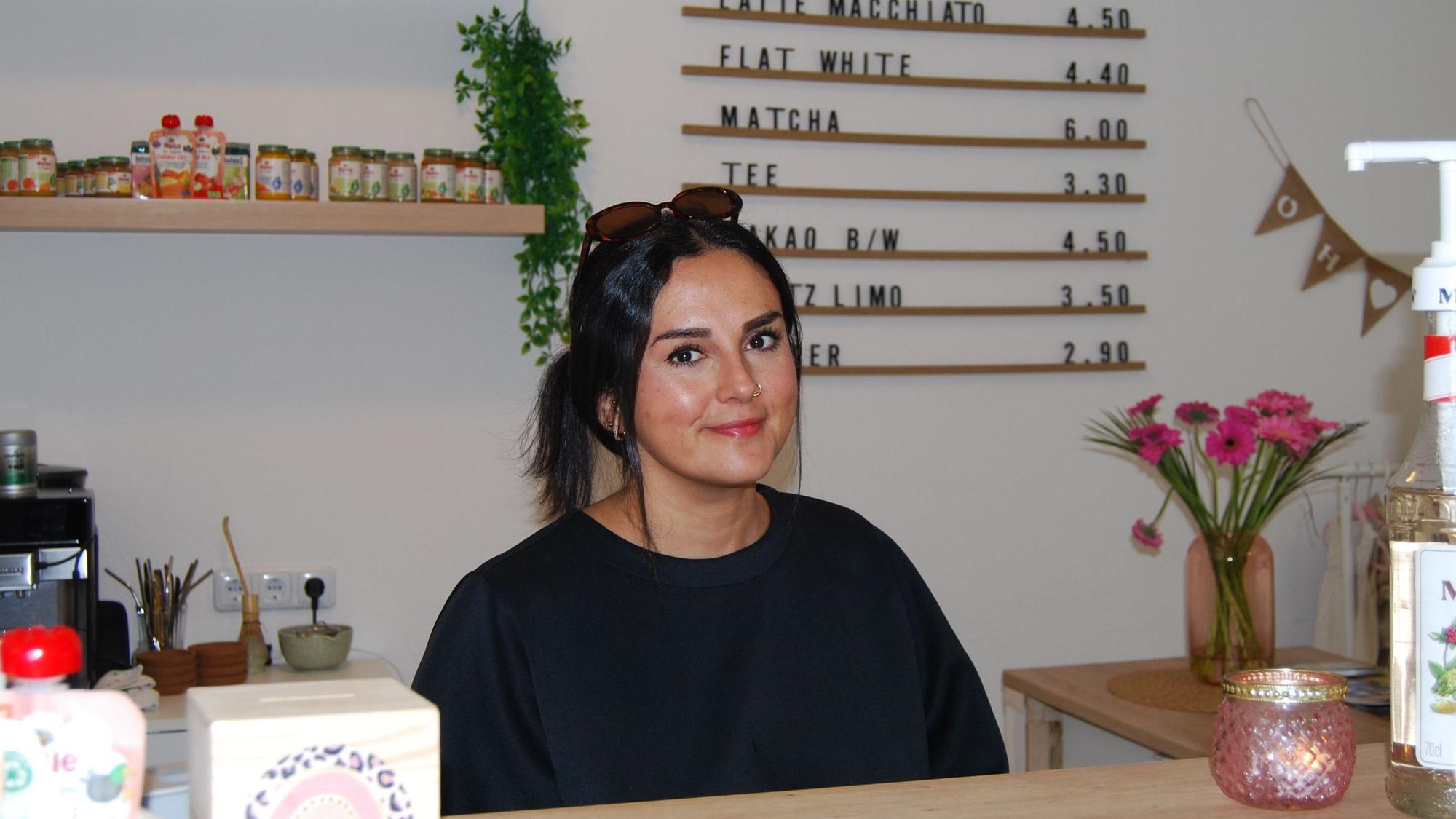 Eine Frau mit dunklen Haaren und Sonnenbrille in den Haaren sitzt hinter einer Theke und lächelt in die Kamera